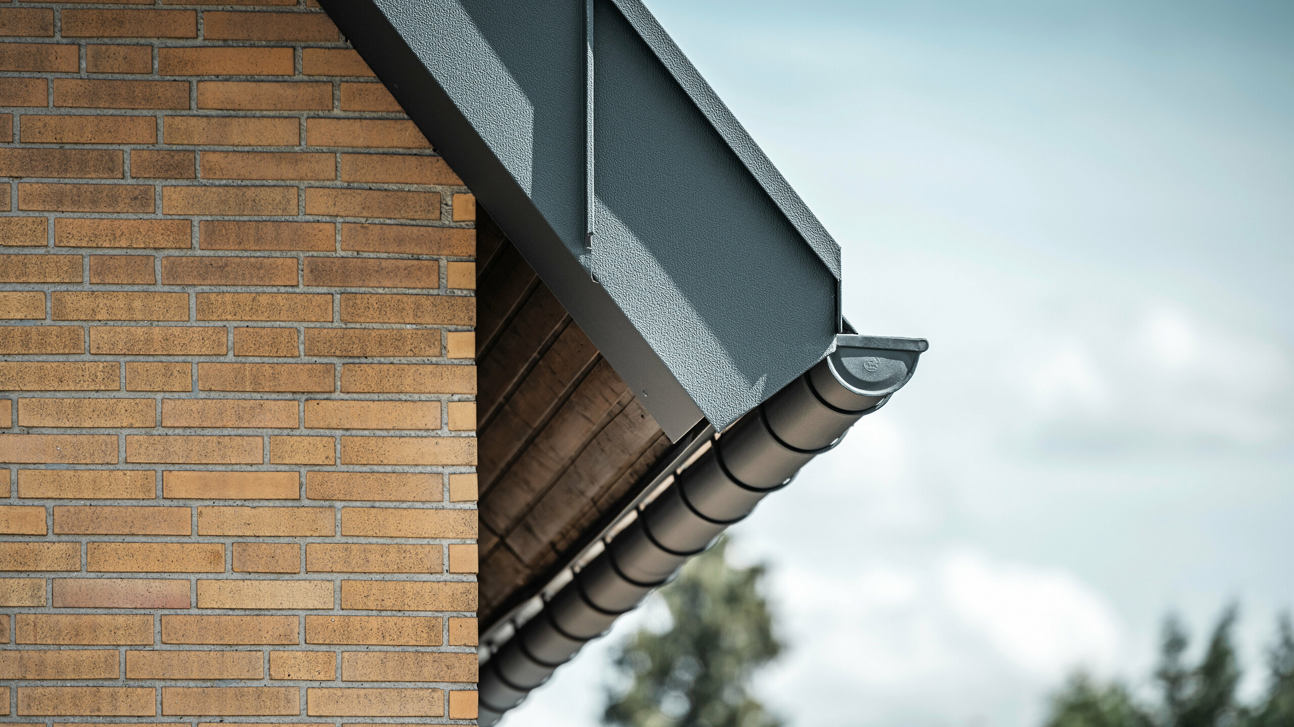 Detailaufnahme eines anthrazitfarbenen Dachentwässerungsystems von PREFA. Die Hängerinne hebt sich deutlich von den warmen Tönen der Backsteinwand ab und betont die präzise Verarbeitung und Qualität der verwendeten Materialien.