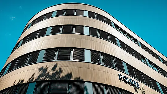 Dramatische Untersicht des geschwungenen Neubaus vom Polizeikommissariat 43 in Hamburg: Die prägnante PREFA Aluminiumfassade aus eloxierten Strangpressprofilen „Zacke“ in Mittelbronze wird zum architektonischen Eyecatcher. Die fein geriffelte Oberfläche erzeugt lebendige Lichtreflexe und betont die fließenden Rundungen sowie die horizontalen Fensterbänder. Dank hoher Korrosionsbeständigkeit, Langlebigkeit und minimalem Wartungsaufwand bieten PREFA Aluminiumlösungen eine nachhaltige, funktionale und ästhetisch anspruchsvolle Fassadengestaltung.