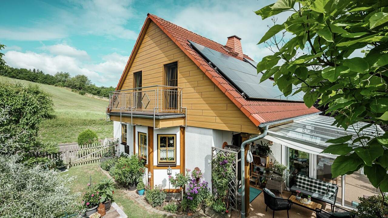 Dieses Bild zeigt ein charmantes Einfamilienhaus in Blieskastel, dessen frisch sanierter Giebel mit PREFA Sidings in Eiche Natur sofort ins Auge fällt. Die warme Holzoptik aus langlebigem Aluminium harmoniert beeindruckend mit dem roten Ziegeldach und dem großzügigen Wintergarten. Umgeben von üppigem Grün und liebevoll angelegten Blumenbeeten entsteht eine idyllische, lebendige Szene. Die robuste PREFA Fassade verleiht dem Haus nicht nur einen natürlichen Look, sondern überzeugt auch durch Witterungsbeständigkeit, Farbtreue und nahezu wartungsfreie Langlebigkeit.