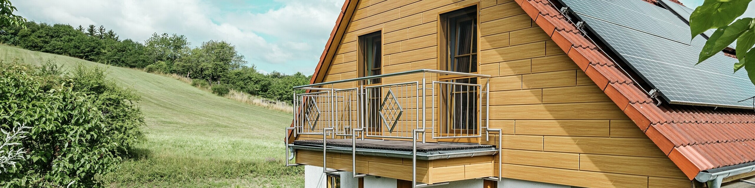 Dieses Bild zeigt ein charmantes Einfamilienhaus in Blieskastel, dessen frisch sanierter Giebel mit PREFA Sidings in Eiche Natur sofort ins Auge fällt. Die warme Holzoptik aus langlebigem Aluminium harmoniert beeindruckend mit dem roten Ziegeldach und dem großzügigen Wintergarten. Umgeben von üppigem Grün und liebevoll angelegten Blumenbeeten entsteht eine idyllische, lebendige Szene. Die robuste PREFA Fassade verleiht dem Haus nicht nur einen natürlichen Look, sondern überzeugt auch durch Witterungsbeständigkeit, Farbtreue und nahezu wartungsfreie Langlebigkeit.