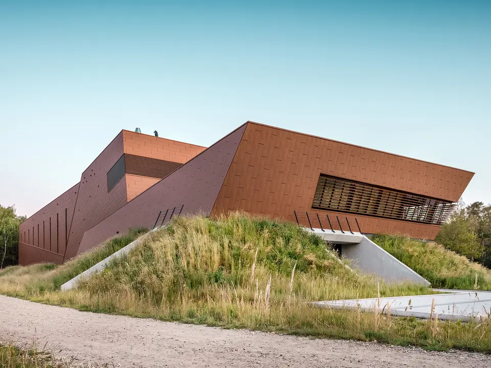 Architektonisch expressives Trinkwasserpumpwerk WML in Lottum mit skulptural gefalteten Baukörpern, eingebettet in eine naturnahe Böschungslandschaft. Die markante PREFA Siding.X Aluminiumfassade in warmer Rostoptik zieht mit ihrer facettierten, lebendigen Oberfläche alle Blicke auf sich und spielt eindrucksvoll mit Licht und Schatten. Horizontale Lamellen und präzise Linienführung betonen die dynamische Geometrie. Die leichten, langlebigen und vollständig recycelbaren PREFA Aluminium-Paneele stehen für höchste Witterungsbeständigkeit, nachhaltige Bauweise und eine wartungsarme Gebäudehülle.