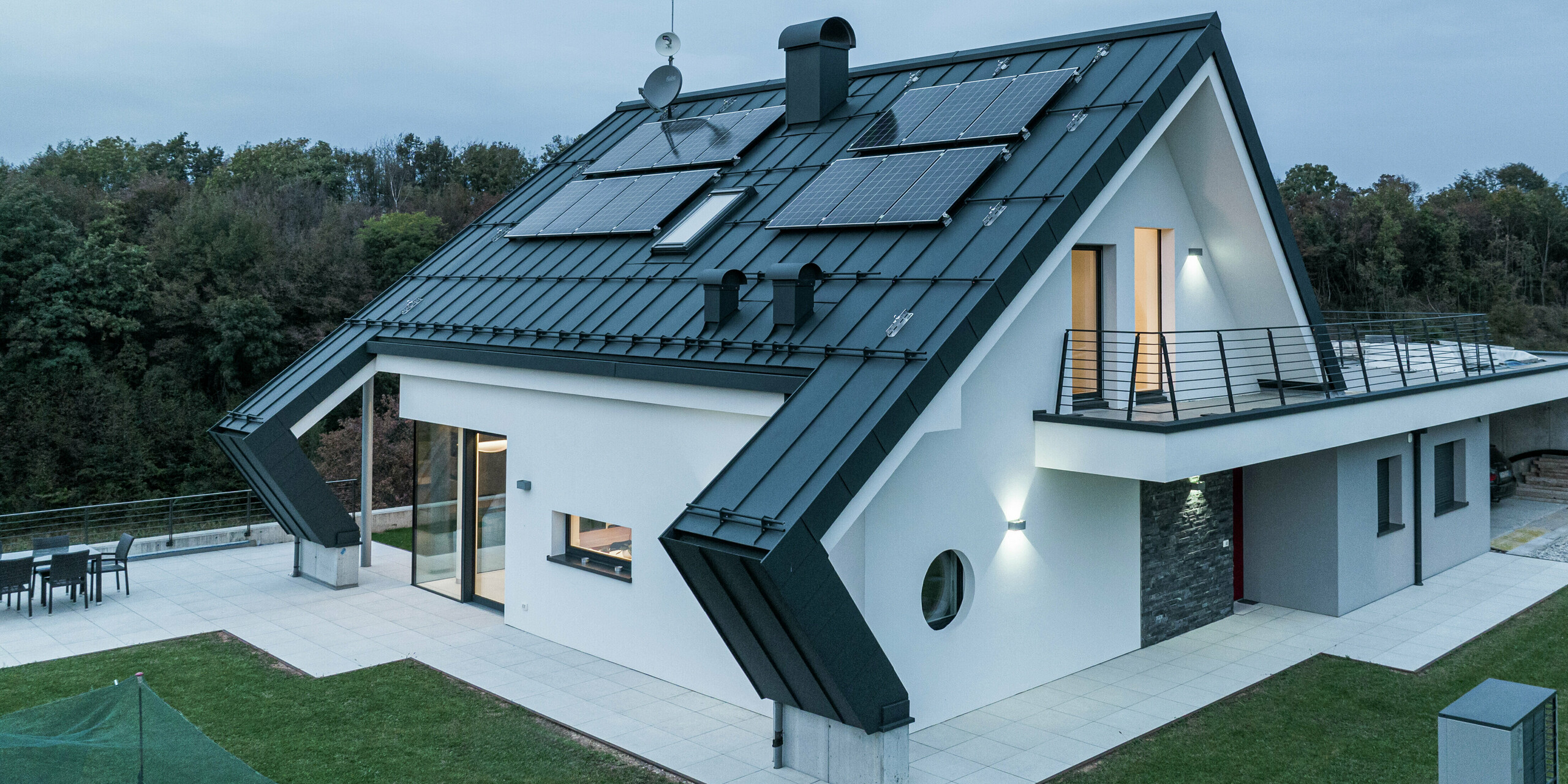 Modernes Einfamilienhaus in Borgo Valbelluna mit PREFALZ Stehfalzdeckung in P.10 Anthrazit. Das Aluminiumdach mit seinen Photovoltaikmodulen verbindet nachhaltige Energiegewinnung mit zeitloser Ästhetik. Die markanten Dachüberstände unterstreichen das innovative Design und bieten zusätzlichen Witterungsschutz. Große Glasflächen, eine weiße Putzfassade und ein umlaufender Balkon ergeben ein harmonisches Gesamtbild.