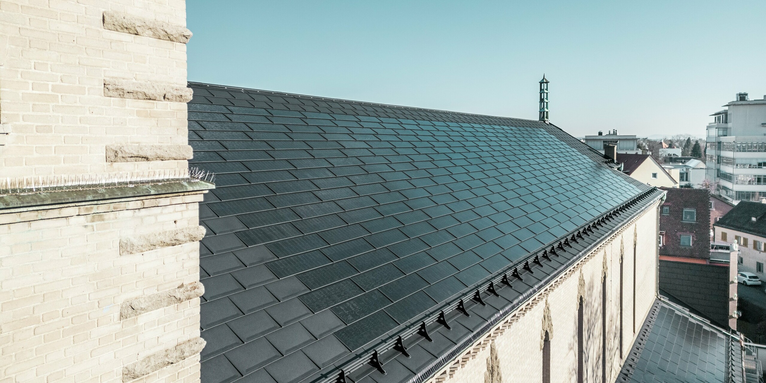 Der Blick am Kirchturm vorbei zeigt die PREFA Solardachplatten in edlem Schwarz auf dem Dach der St. Karl Borromäus Kirche in Nürnberg. Die Perspektive verdeutlicht die perfekte Integration der Solarmodule, die sich nahtlos in das Satteldach des denkmalgeschützten Gebäudes einfügen. Auch die prägnanten Zierelemente der Kirchenmauer, welche die Architektur des fränkischen Expressionismus unterstreichen, sind erkennbar. Die Solardachplatten liefern nachhaltige Energie für die Kirche und die umliegenden Gebäude und stellen gleichzeitig eine ästhetische Lösung im Bereich des Denkmalschutzes dar.