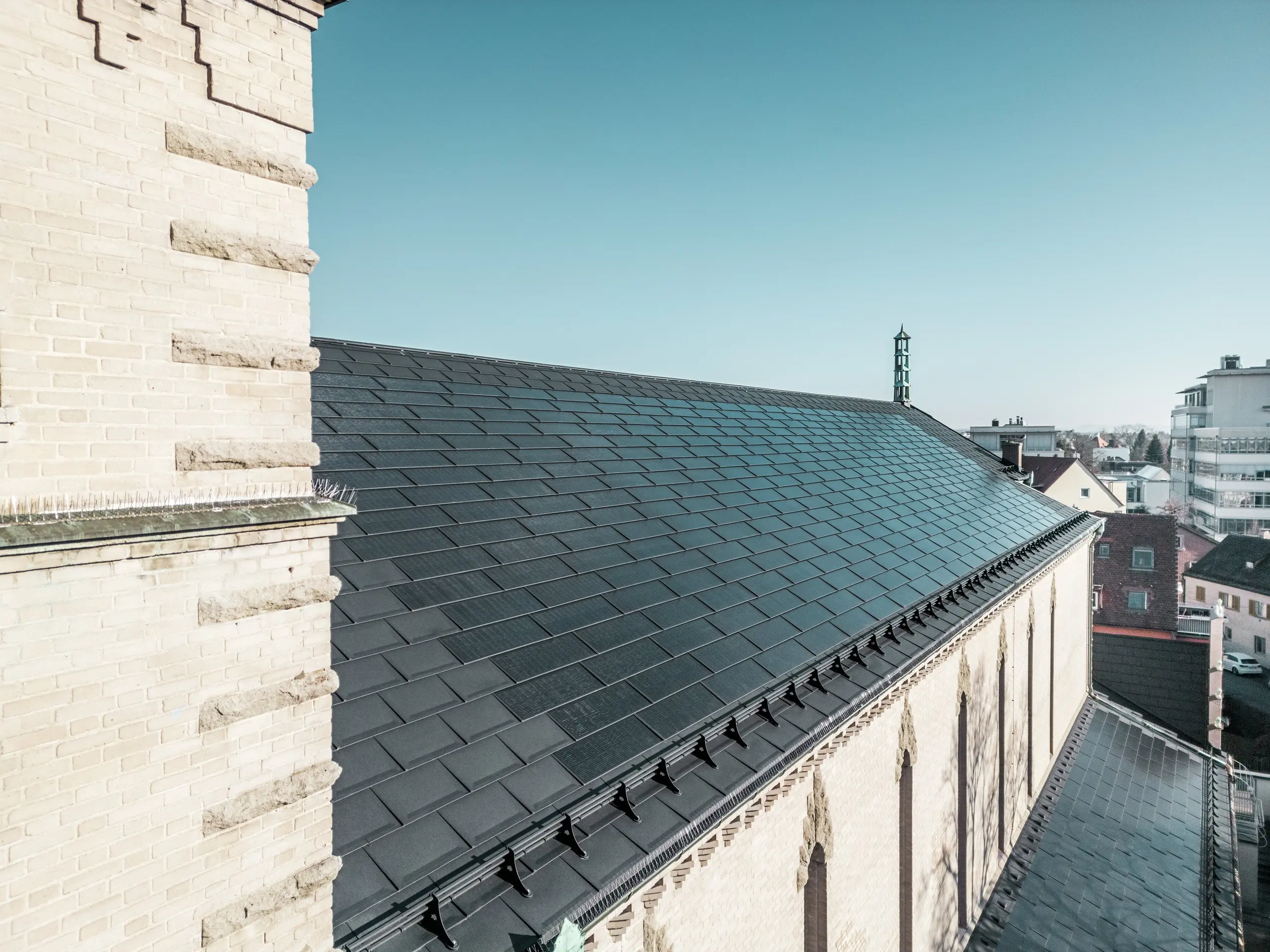 Der Blick am Kirchturm vorbei zeigt die PREFA Solardachplatten in edlem Schwarz auf dem Dach der St. Karl Borromäus Kirche in Nürnberg. Die Perspektive verdeutlicht die perfekte Integration der Solarmodule, die sich nahtlos in das Satteldach des denkmalgeschützten Gebäudes einfügen. Auch die prägnanten Zierelemente der Kirchenmauer, welche die Architektur des fränkischen Expressionismus unterstreichen, sind erkennbar. Die Solardachplatten liefern nachhaltige Energie für die Kirche und die umliegenden Gebäude und stellen gleichzeitig eine ästhetische Lösung im Bereich des Denkmalschutzes dar.