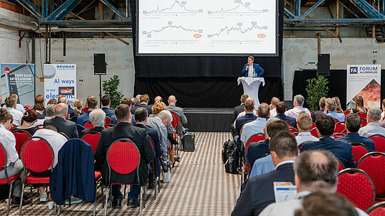 Großer Konferenzsaal im industriellen Ambiente des Glanzstoff-Geländes mit dicht besetzten Reihen und roter Bestuhlung als visuellem Kontrast. Im Fokus steht eine beleuchtete Bühne mit Rednerpult und Präsentationsfläche, auf der detaillierte Marktanalysen zu Rohstoff- und Energiepreisen gezeigt werden. Flankiert von Ausstellerbannern wie PREFA Dachentwässerung und Neuman Aluminium entsteht eine dynamische Atmosphäre. Das Bild vermittelt den intensiven Austausch führender Experten beim Forum Aluminium zu Innovationen, Markttrends und Zukunft der Branche.