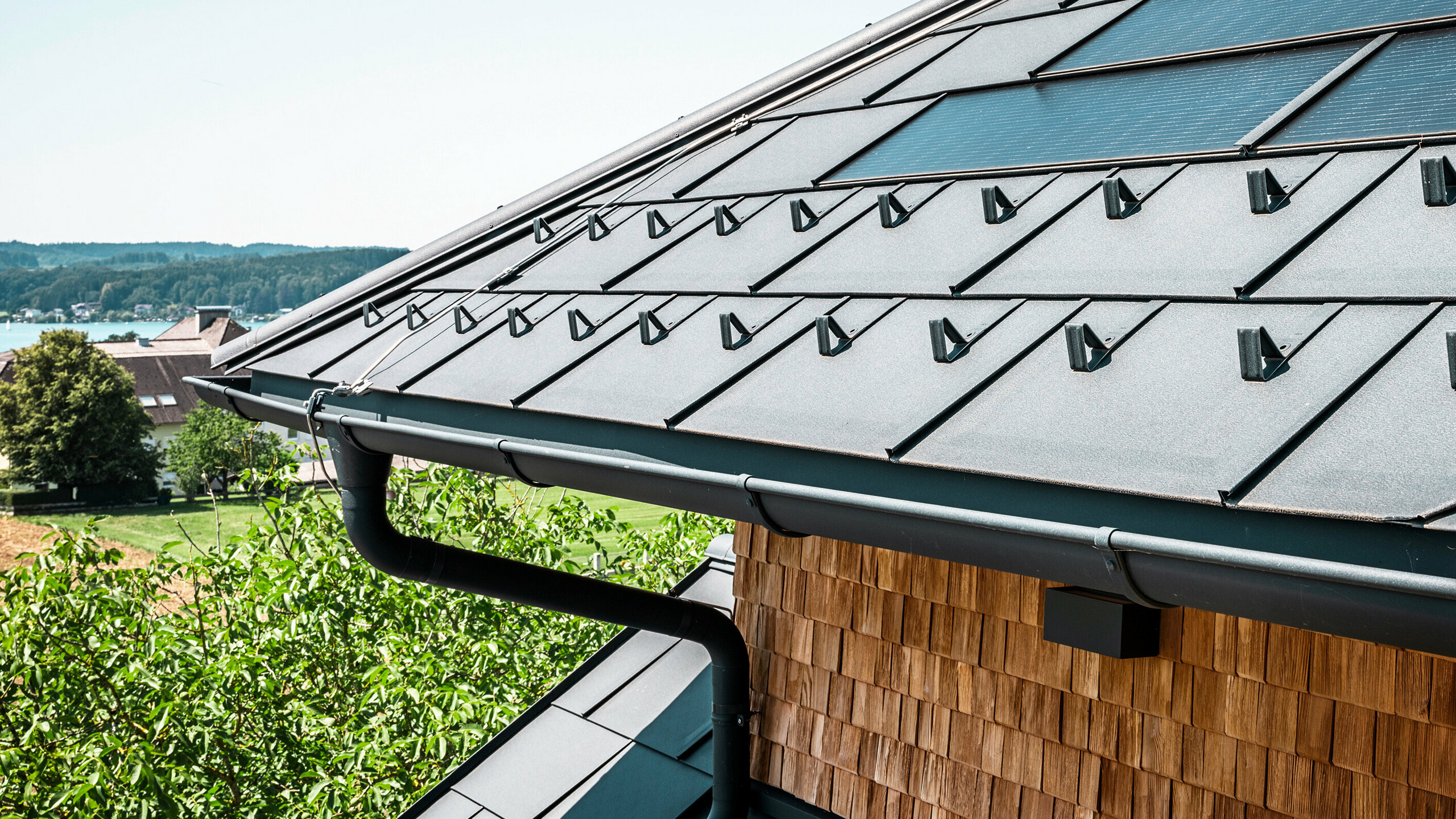 Die Zenz'n Stub'n in Schörfling am Attersee präsentiert ein neu saniertes Dach mit innovativen PREFA Solardachplatten und langlebigen PREFA Dachplatten R.16 in P.10 Anthrazit. Das stilvolle Aluminiumdach verbindet modernes Design mit nachhaltiger Energiegewinnung. Auch die Dachentwässerung, bestehend aus Ablaufrohr und Hängerinne in P.10 Anthrazit, fügt sich harmonisch in das Gesamtbild ein und unterstreicht die hochwertige Verarbeitung. Im Hintergrund blitzt das Blau des Attersees hervor.
