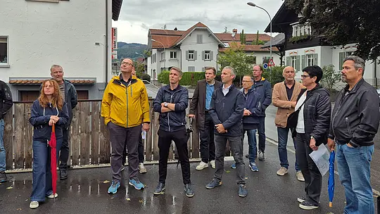 Teilnehmende der PREFA Objekttour stehen auf einer regennassen Straße und blicken aufmerksam zur vorgestellten Dachlösung. Der leuchtend gelbe Regenmantel setzt einen markanten Farbakzent im grauen Wetter. Im Fokus stehen die hochwertige Architektur sowie die langlebigen und wetterbeständigen PREFA Aluminiumlösungen für Dach und Gebäudehülle.