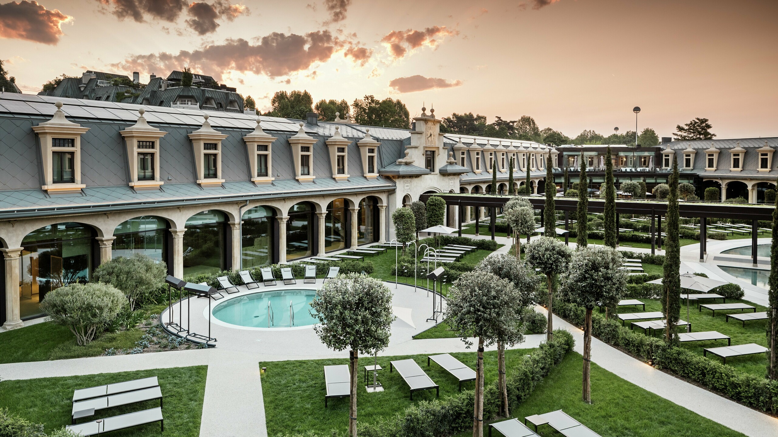 Abendstimmung im Thermalpark „De Montel – Terme Milano“ mit eindrucksvollem Blick auf die denkmalgeschützte Fassadenarchitektur im Jugendstil. Die Dacheindeckung mit PREFA Dachrauten 44 × 44 in P.10 Hellgrau verbindet Nachhaltigkeit mit Ästhetik. Im Vordergrund laden Liegewiesen, gepflegte Wege, Olivenbäume und ein rundes Thermalbecken zum Verweilen ein.