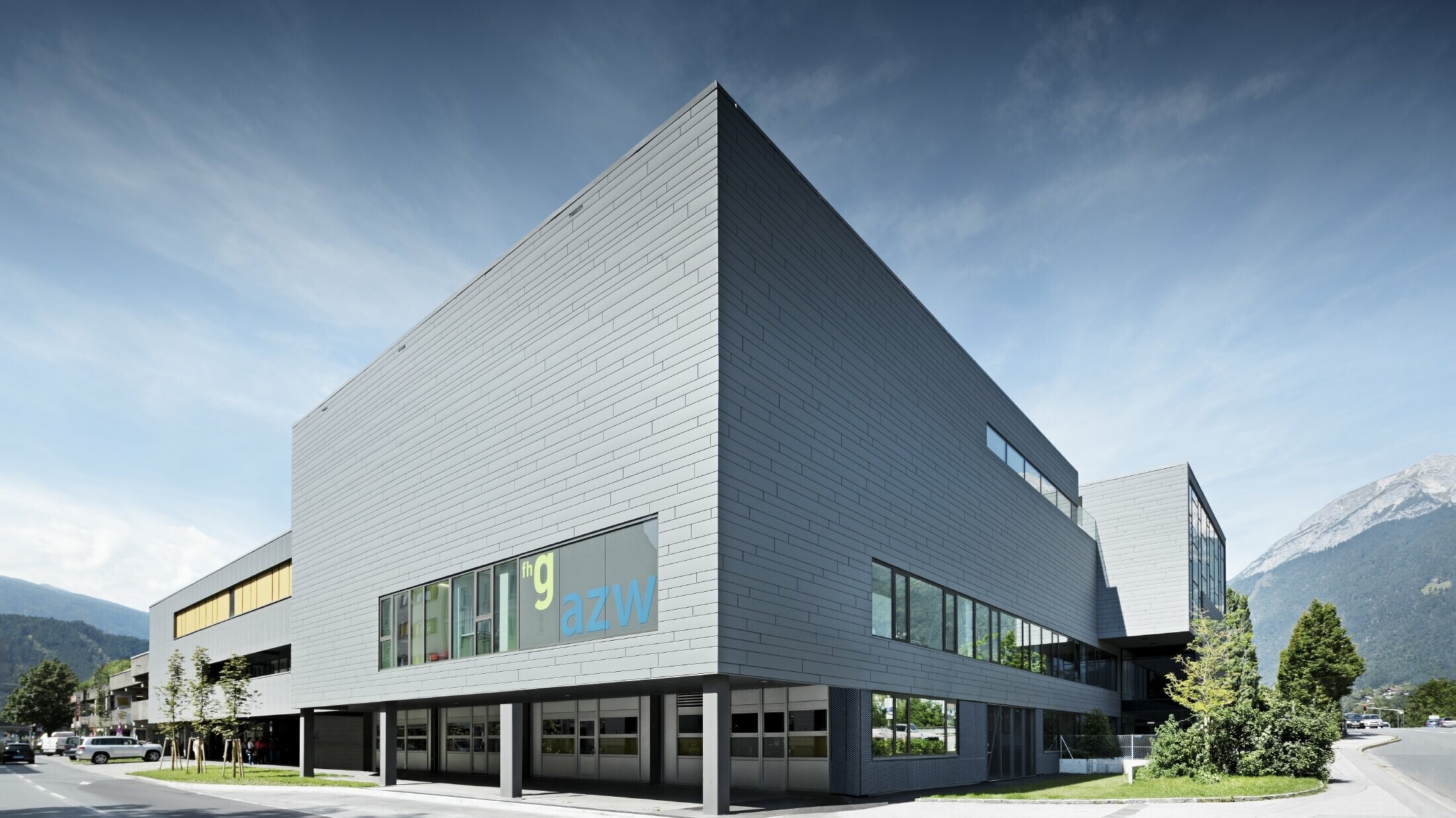 Modernes Ausbildungszentrum mit klarer, kubischer Architektur und hellgrauer Aluminiumfassade. Das Gebäude hat große Fensterflächen und überhängende Baukörper. Auf einer Glasfront ist die Beschriftung „fhg azw“ zu sehen. Im Hintergrund erheben sich Berge unter einem leicht bewölkten Himmel, während im Vordergrund eine gepflegte Straße mit Grünflächen verläuft.