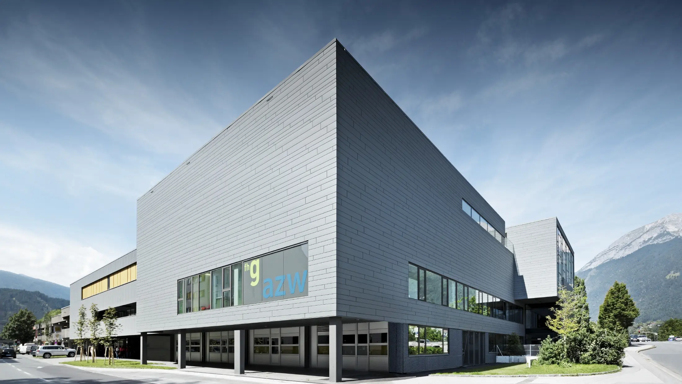 Modernes Ausbildungszentrum mit klarer, kubischer Architektur und hellgrauer Aluminiumfassade. Das Gebäude hat große Fensterflächen und überhängende Baukörper. Auf einer Glasfront ist die Beschriftung „fhg azw“ zu sehen. Im Hintergrund erheben sich Berge unter einem leicht bewölkten Himmel, während im Vordergrund eine gepflegte Straße mit Grünflächen verläuft.