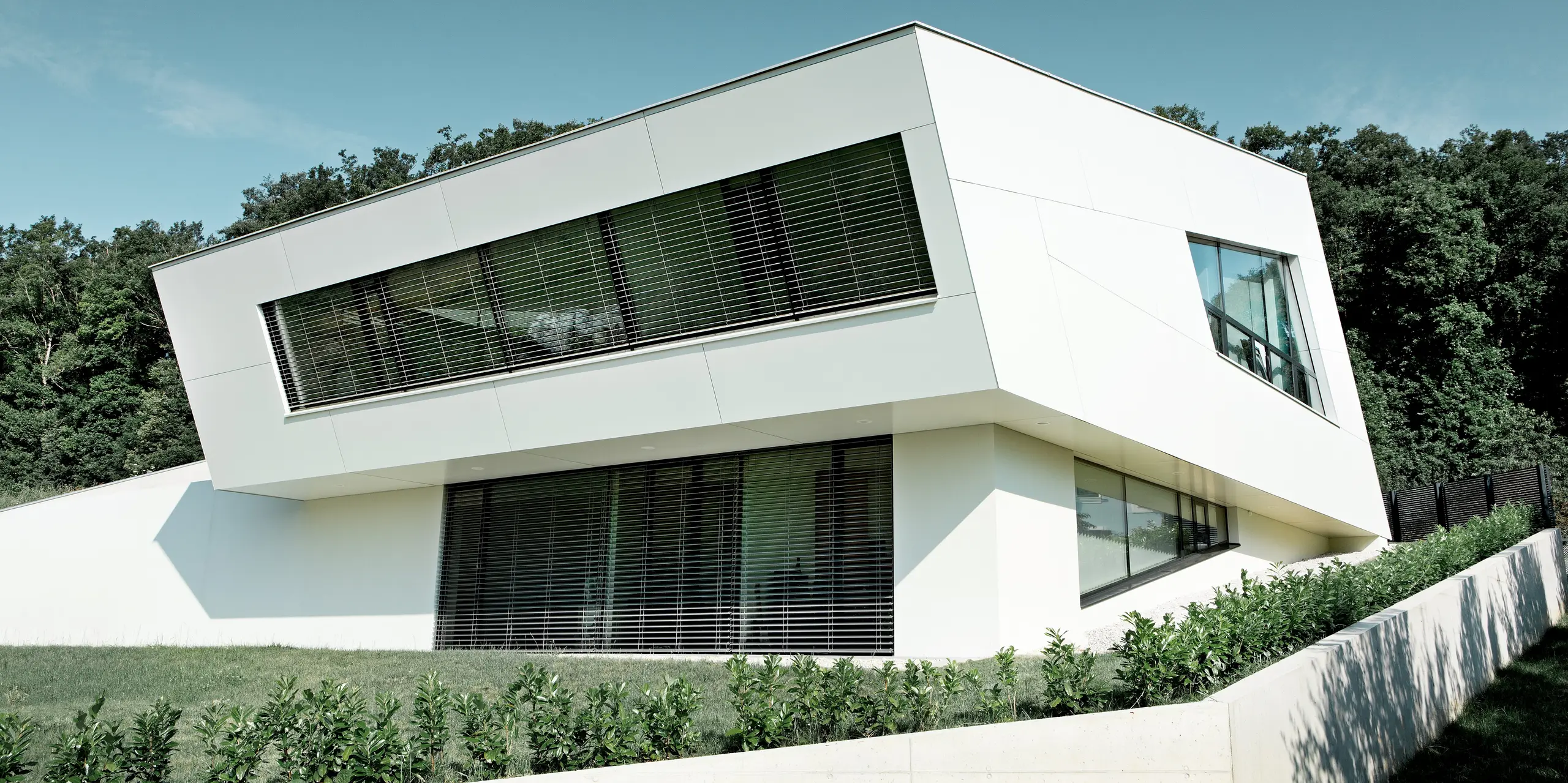 Modernes Einfamilienhaus in Bad Tatzmannsdorf mit kubischer Architektur, großen Fenstern und weißen Fassadenplatten; davor gepflegter Rasen und kleine Hecken, blauer Himmel und Bäume im Hintergrund.