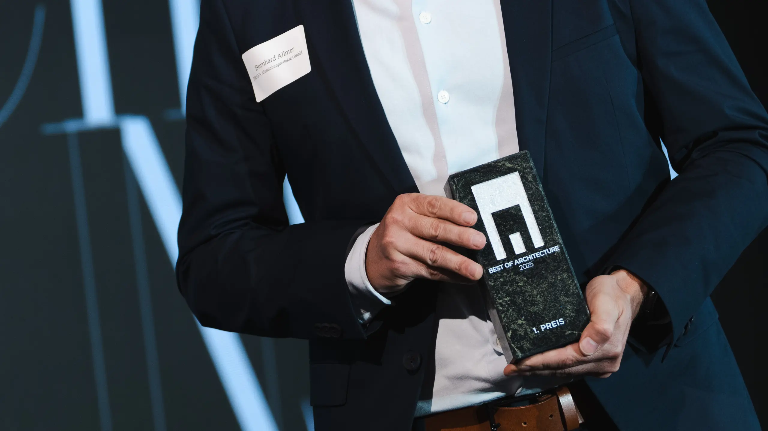 Die Preisverleihung beim renommierten Callwey-Wettbewerb „Wohngebäude des Jahres“ 2025: Im Vordergrund präsentiert eine elegante Gestalt in dunklem Anzug und weißem Hemd stolz den Preis „Architects' Choice“. Die Person ist Bernhard Allmer, der für PREFA-Solarprodukte verantwortlich ist. Der Preis besteht aus einer eleganten dunklen Steinplatte mit einem modernen Logo und klarer Typografie. Im Hintergrund befindet sich eine stilvolle Bühne mit einem großen Banner und Akzentbeleuchtung, die den feierlichen Charakter der Veranstaltung unterstreicht. Die Szene spiegelt Innovation und Exzellenz in der Architektur wider.
