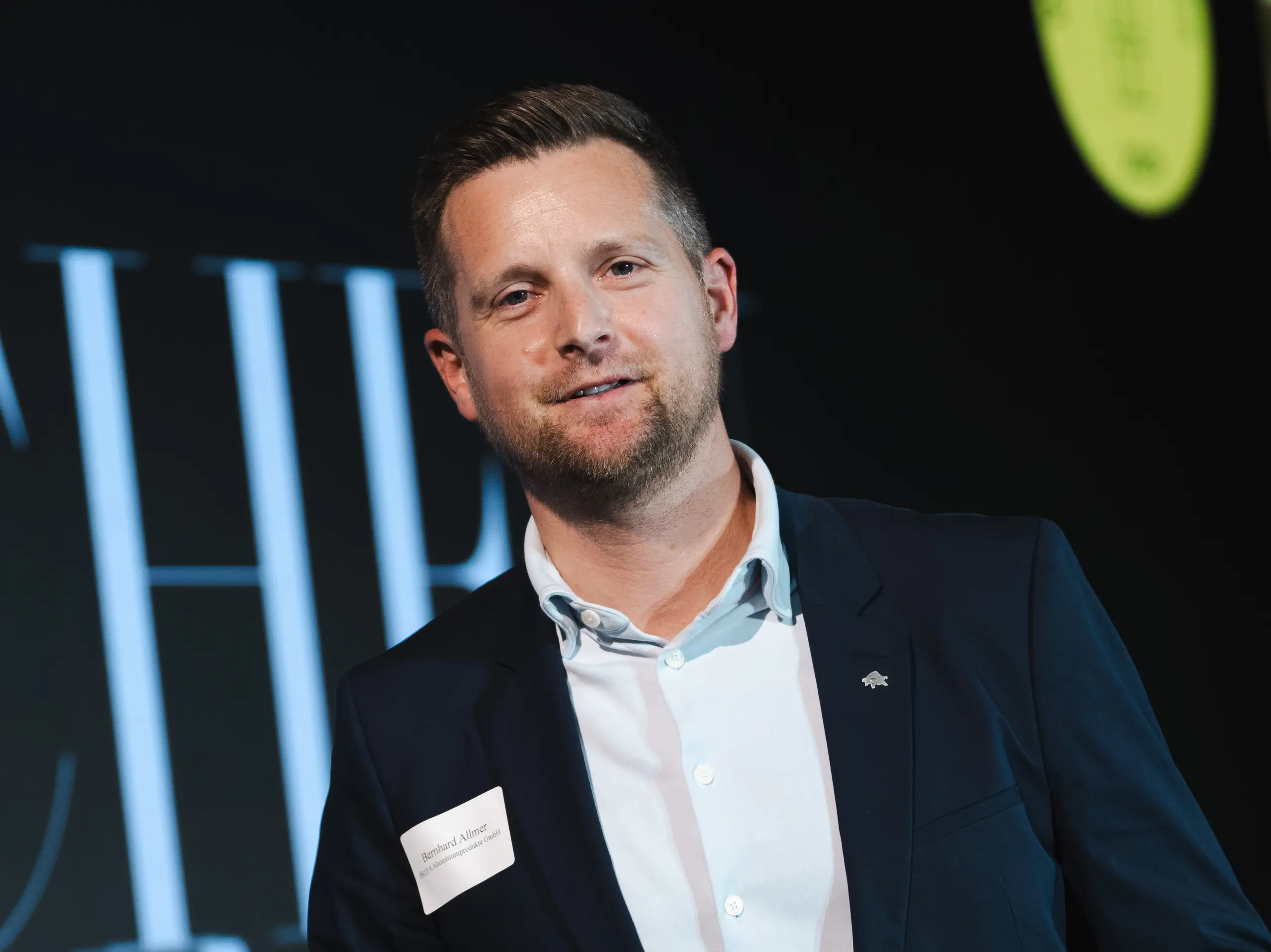 Die Preisverleihung beim renommierten Callwey-Wettbewerb „Wohngebäude des Jahres“ 2025: Im Vordergrund präsentiert eine elegante Gestalt in dunklem Anzug und weißem Hemd stolz den Preis „Architects' Choice“. Die Person ist Bernhard Allmer, der für PREFA-Solarprodukte verantwortlich ist. Der Preis besteht aus einer eleganten dunklen Steinplatte mit einem modernen Logo und klarer Typografie. Im Hintergrund befindet sich eine stilvolle Bühne mit einem großen Banner und Akzentbeleuchtung, die den feierlichen Charakter der Veranstaltung unterstreicht. Die Szene spiegelt Innovation und Exzellenz in der Architektur wider.
