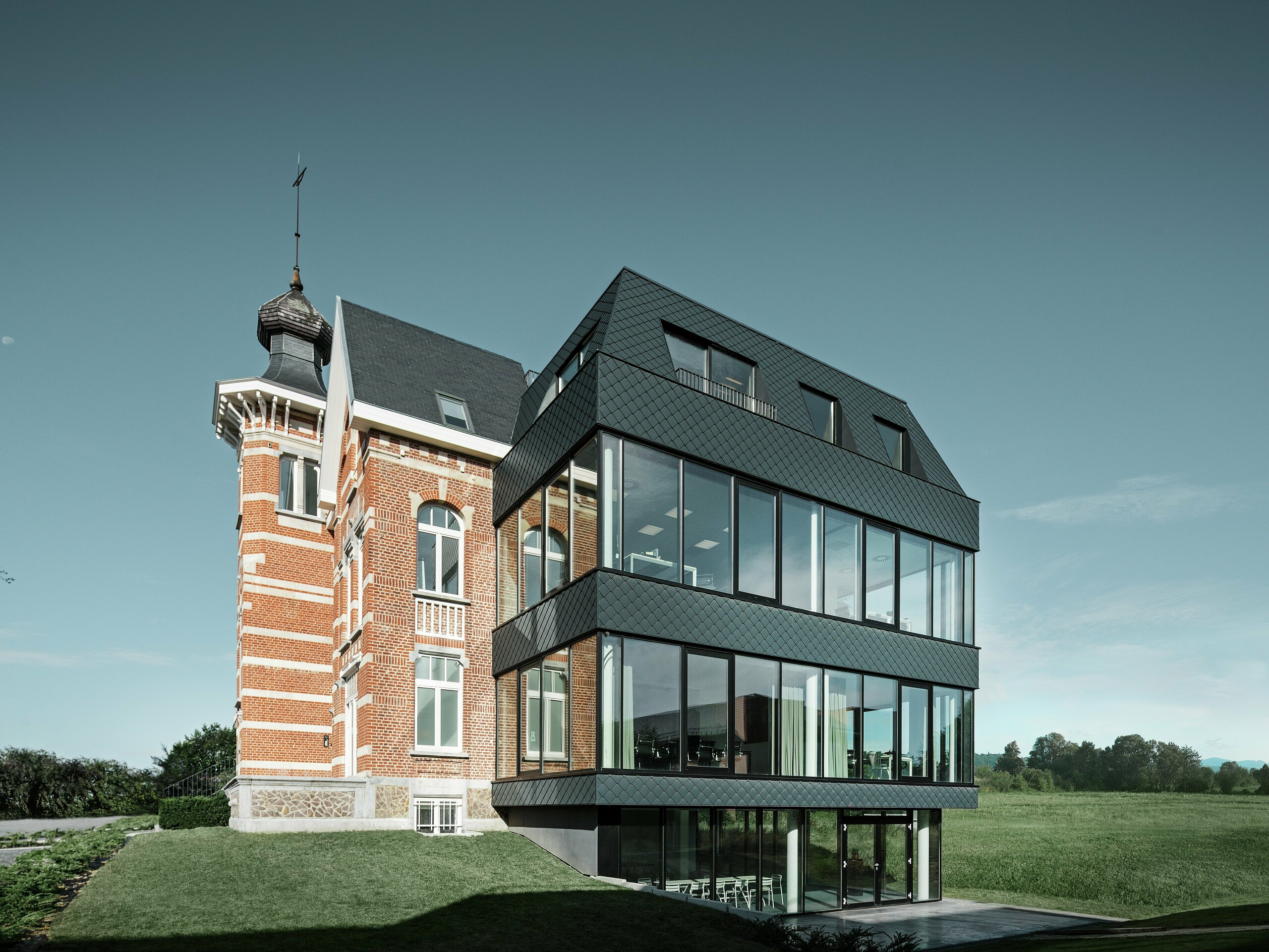 Seitlicher Blick auf das Kasteeltje Heizel, wo historischer Backsteinbau mit Turm nahtlos in einen modernen Anbau übergeht. Die anthrazitfarbenen PREFA Wandrauten setzen mit ihrer markanten Struktur elegante Kontraste zu den großzügigen Glasflächen. Die klare Linienführung des Neubaus verstärkt die Verbindung von Tradition und moderner Architektur in einer eindrucksvollen Kulisse.