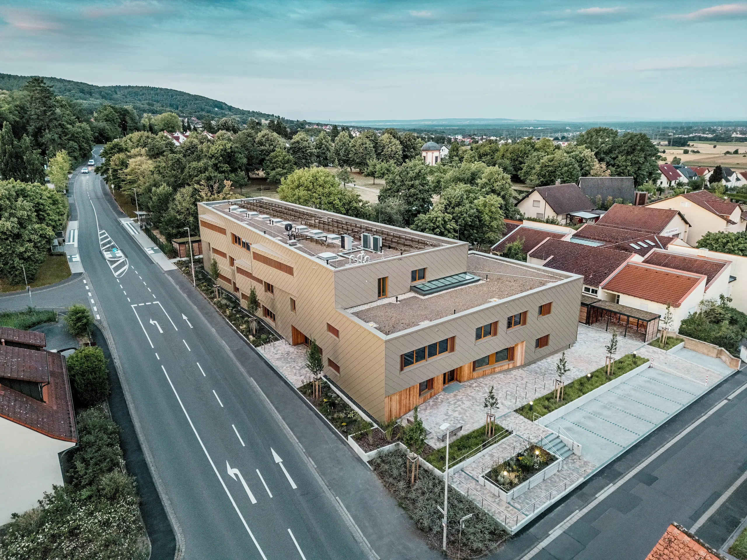 Die eindrucksvolle Gesamtgestaltung der kommunalen Kindertagesstätte in Alzenau wird auf der Luftaufnahme sichtbar. Das winkelförmige Hybridgebäude ist harmonisch in ein ruhiges Wohnviertel am Rande des Spessarts eingebettet. Die Fassade aus bronzefarbenen PREFA Wandrauten 44 × 44 verleiht dem Baukörper eine markante, hochwertige Optik und betont die moderne Architektur. Begrünte Dachflächen, großzügige Außenanlagen und eine klare Verkehrsführung unterstreichen das nachhaltige Gesamtkonzept. Die Kombination aus langlebigem Aluminium, Holz und Recyclingbeton erfüllt höchste ökologische Standards. Diese Perspektive hebt die durchdachte städtebauliche Integration und den multifunktionalen Charakter des Gebäudes hervor – ein zukunftsweisendes Projekt für Bildung, Wohnen und Nachhaltigkeit.