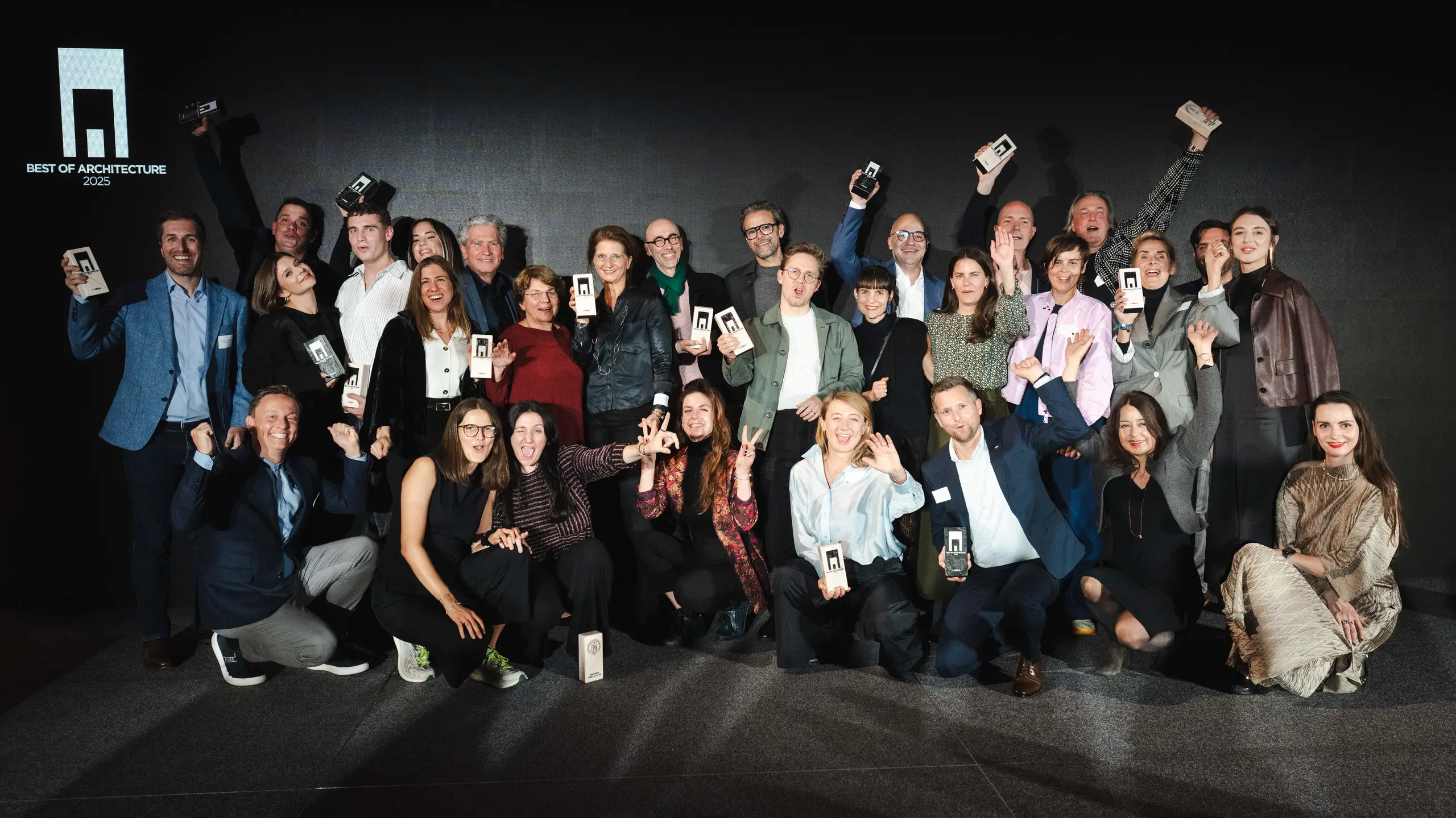 Gruppenfoto der Gewinner bei der Preisverleihung „Wohngebäude des Jahres 2025“: Die Gewinner stehen vor einer eleganten schwarzen Bühne mit dem Logo „Best of Architecture“ und halten stolz ihre Auszeichnungen in den Händen. Dynamische Posen, moderne Kleidung und auffällige Beleuchtung unterstreichen die Bedeutung der Veranstaltung, während die Bühne für Innovation und architektonische Exzellenz steht.