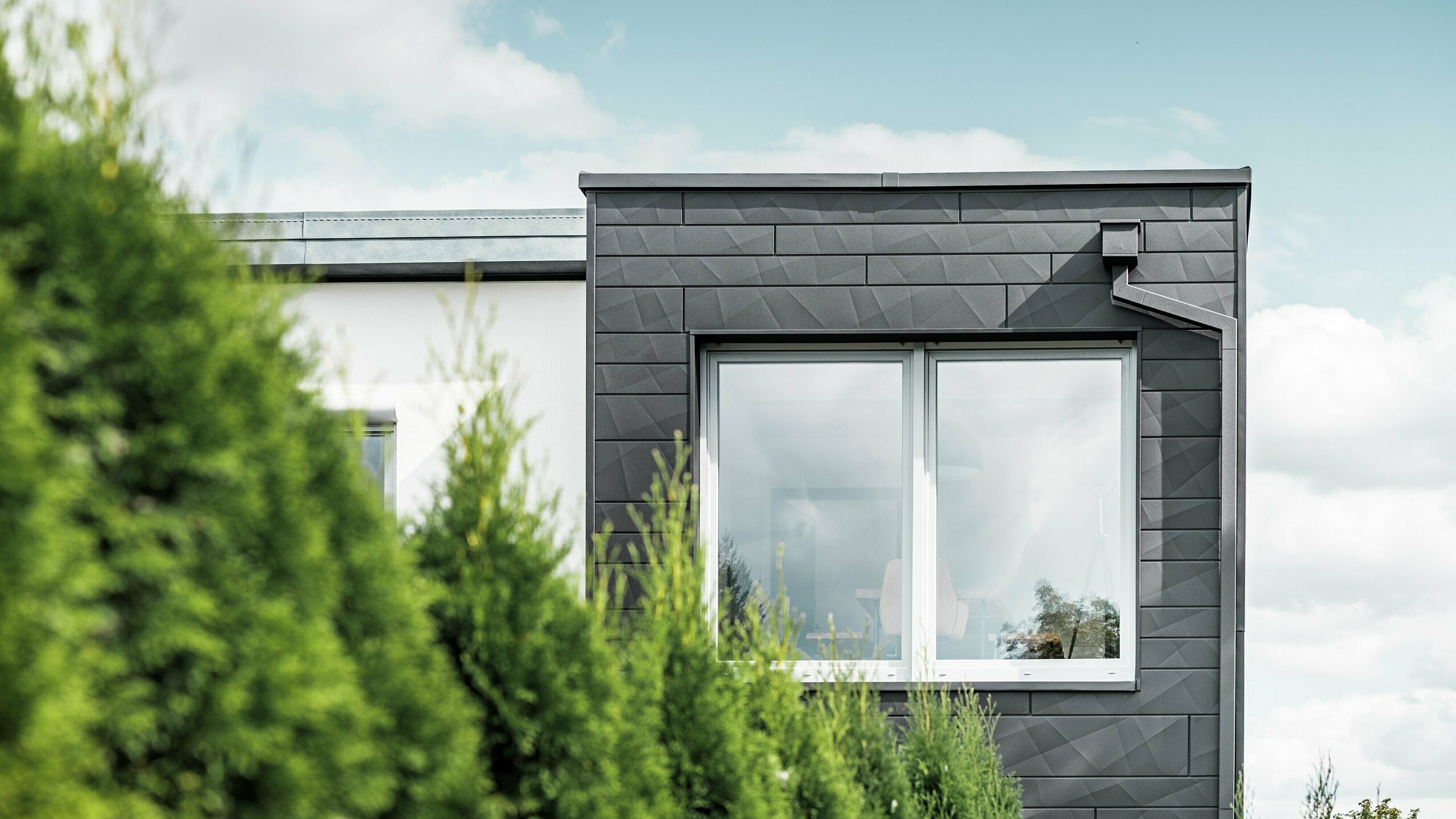 Die klar strukturierten PREFA Siding.X Fassadenpaneele in der Farbe P.10 Schwarz zieren die Außenwand eines Einfamilienhauses in Teuschnitz und verleihen ihm ein einzigartiges Erscheinungsbild. Die PREFA Dachentwässerung, bestehend aus dem Quadratrohr und dem Wasserfangkasten, fügt sich nahtlos in das Design ein. Die grünen Thuja-Bäume im Vordergrund kontrastieren mit der dunklen Blechfassade.
