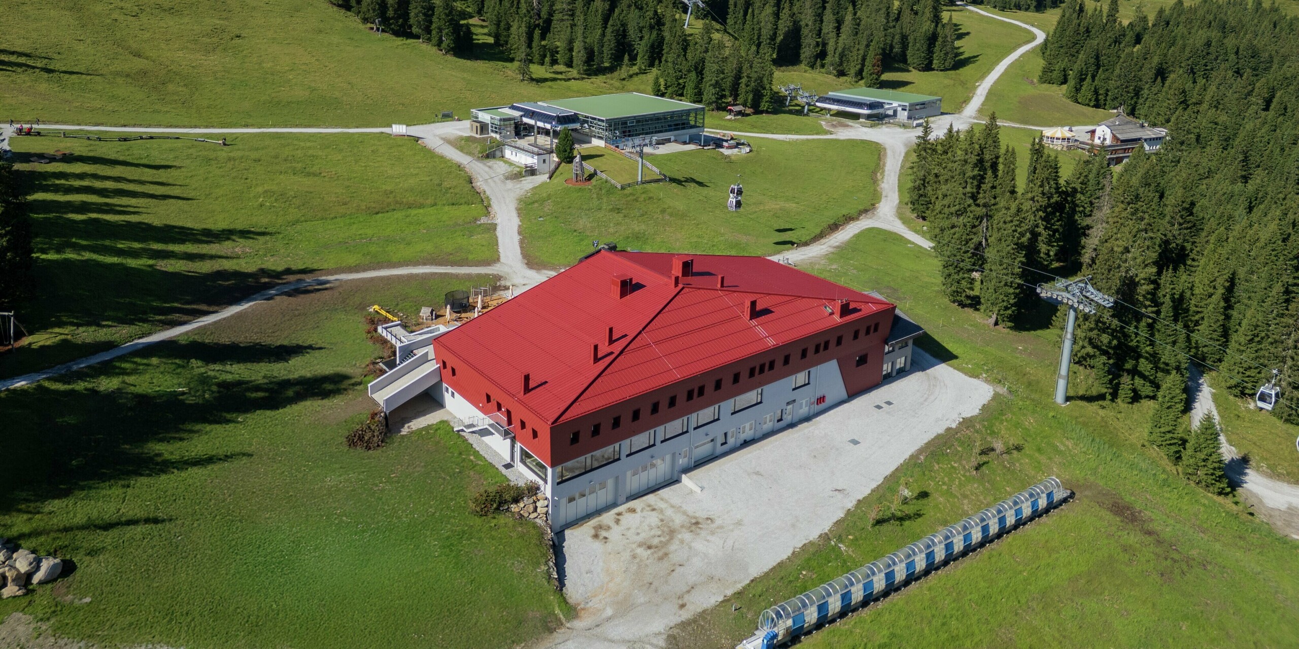 Das beeindruckende Gebäude des Summit Mountain Club in Ratschings hebt sich markant von der alpinen Landschaft ab. Besonders auffällig ist die oxydrote Aluminiumhülle mit PREFALZ am Dach und PREFA Wandschindeln an der Fassade. Im Vordergrund sind großzügige Wiesenflächen und ein Zufahrtsweg zum Gebäude zu erkennen. Die Liftstation und das weitläufige Gelände im Hintergrund betonen die Bergwelt, während das Gebäude durch seine moderne und funktionale Gestaltung sowohl als Restaurant als auch als Veranstaltungsort dient.