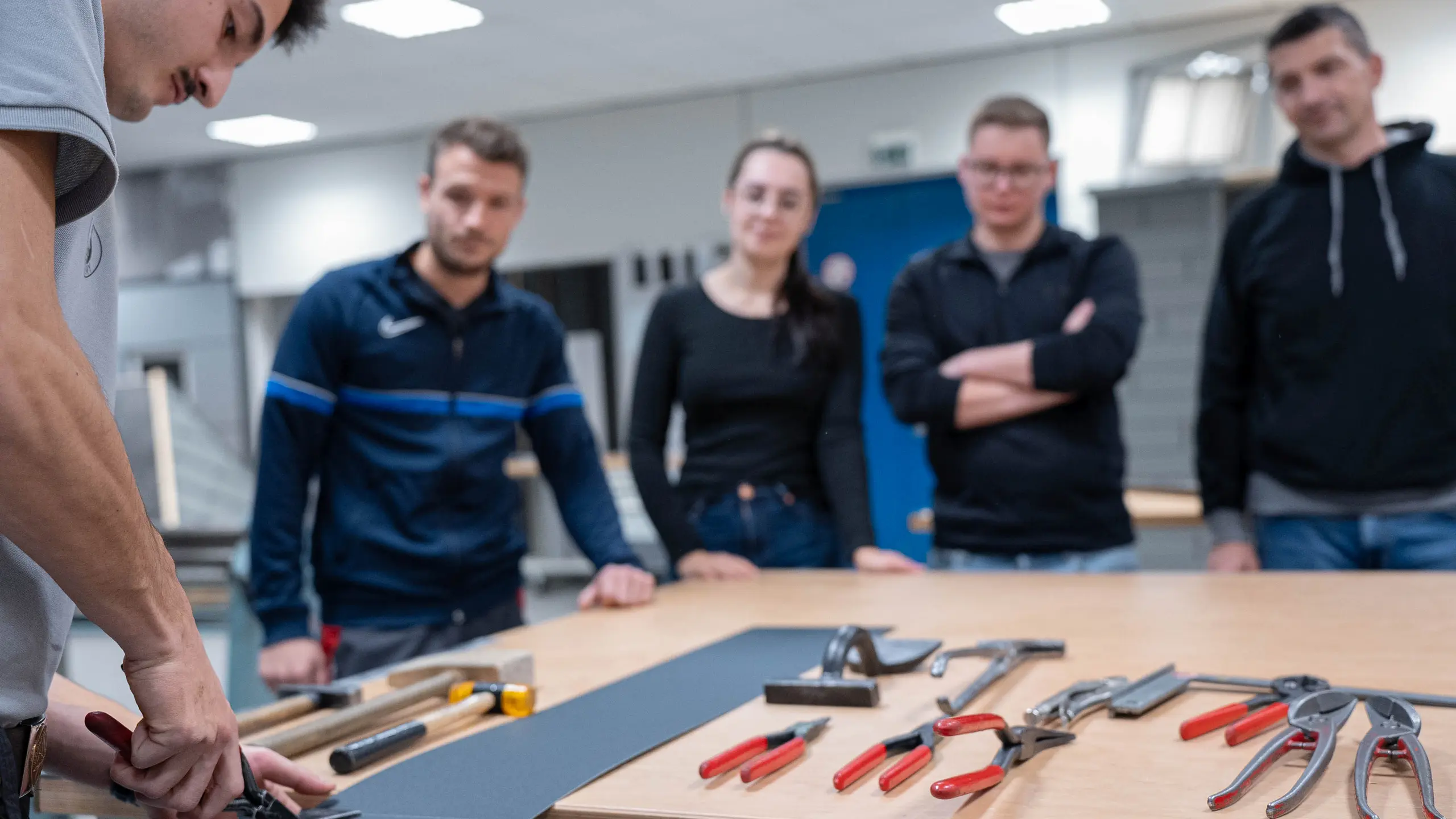 Praktische Schulungssituation in der PREFA Academy: Ein Ausbilder demonstriert die Handhabung von Werkzeugen auf einem Tisch, während die Teilnehmer aufmerksam zuhören und lernen. Das Bild steht für die praxisorientierten Weiterbildungsmöglichkeiten bei PREFA, die Handwerkern und Fachleuten wertvolle Fähigkeiten im Umgang mit PREFA-Produkten vermitteln.