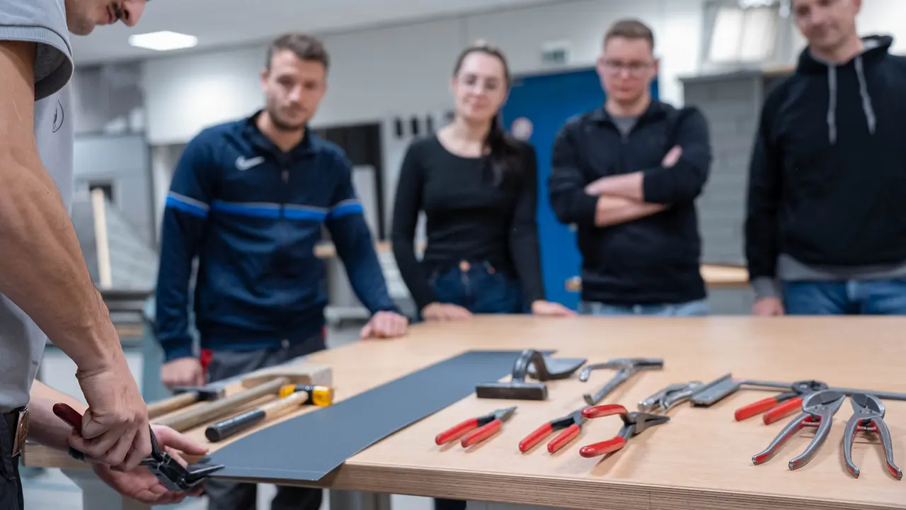 Praktische Schulungssituation in der PREFA Academy: Ein Ausbilder demonstriert die Handhabung von Werkzeugen auf einem Tisch, während die Teilnehmer aufmerksam zuhören und lernen. Das Bild steht für die praxisorientierten Weiterbildungsmöglichkeiten bei PREFA, die Handwerkern und Fachleuten wertvolle Fähigkeiten im Umgang mit PREFA-Produkten vermitteln.