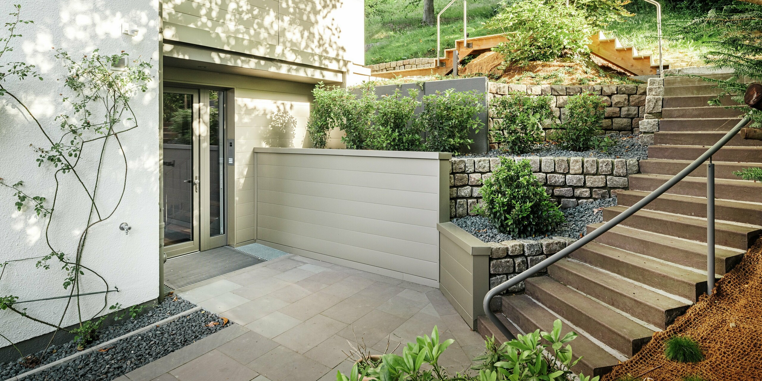 Der Eingangsbereich der Villa Blankenese in Hamburg zeigt, wie moderne Architektur und hochwertige Materialien harmonisch zusammenspielen. Die klare Linienführung und die Kombination aus Naturstein, Grünflächen und Metall schaffen ein elegantes Gesamtbild. Besonders auffällig sind die bronzefarbenen Fassadenelemente aus PREFA Sidings, die dem Objekt einen warmen und edlen Charakter verleihen.