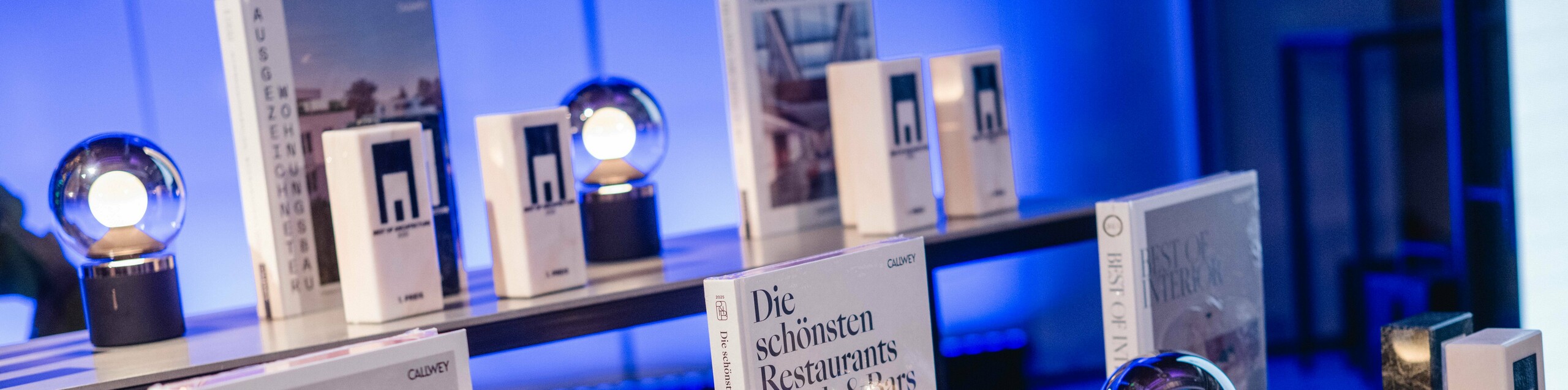 Ausstellung der Awards beim Wettbewerb ‚Wohnbauten des Jahres‘ 2025: Mehrere elegante Trophäen mit dem Logo ‚Best of Architecture‘ stehen auf einem modernen Tisch, flankiert von hochwertigen Architektur- und Designbüchern wie ‚Best Workspaces‘ und ‚Die schönsten Restaurants Hotels & Bars‘. Die Szene ist in stimmungsvolles blaues Licht getaucht und vermittelt Exklusivität, Innovation und architektonische Spitzenleistungen.