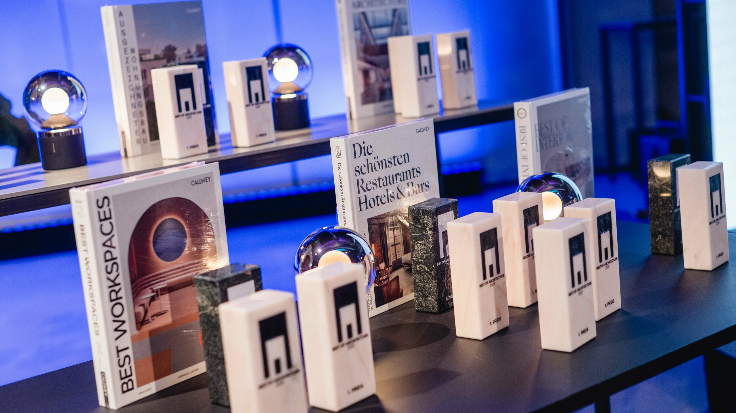 Ausstellung der Awards beim Wettbewerb ‚Wohnbauten des Jahres‘ 2025: Mehrere elegante Trophäen mit dem Logo ‚Best of Architecture‘ stehen auf einem modernen Tisch, flankiert von hochwertigen Architektur- und Designbüchern wie ‚Best Workspaces‘ und ‚Die schönsten Restaurants Hotels & Bars‘. Die Szene ist in stimmungsvolles blaues Licht getaucht und vermittelt Exklusivität, Innovation und architektonische Spitzenleistungen.