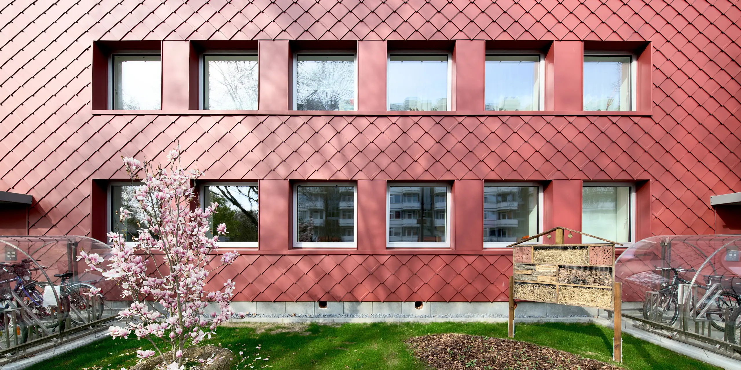 Markante PREFA Wandrauten 29 × 29 in P.10 Oxydrot verwandeln das sanierte Mehrfamilienhaus „Energiesprong 2426“ in Köln in einen architektonischen Blickfang. Das lebendige Rautenmuster mit changierenden Oberflächen, moderne Fensterbänder und ein blühender Magnolienbaum schaffen spannende Kontraste. Die leichte, langlebige und witterungsbeständige Aluminiumfassade vereint Ästhetik, Nachhaltigkeit und Wartungsarmut.