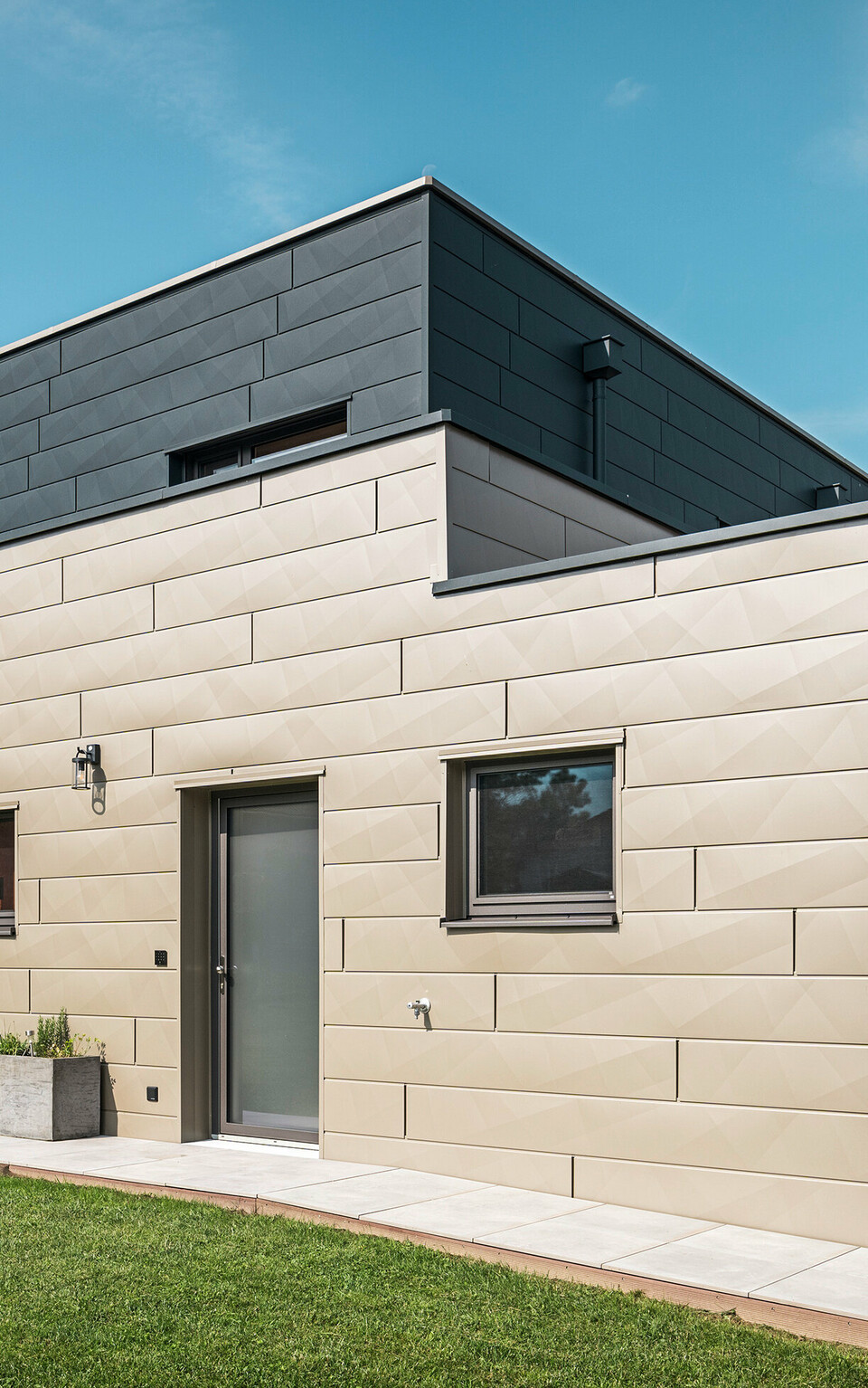 Modernes Einfamilienhaus in Wippenham, Österreich, mit zweifarbiger PREFA Aluminiumfassade in Bronze und Anthrazit. Die Fassade ist mit Siding.X Fassadenpaneelen gestaltet, die für eine elegante und zeitgemäße Optik sorgen. Die integrierte PREFA Dachentwässerung mit Wasserfangkasten und Ablaufrohr sowie eine gepflegte Gartenanlage mit Terrasse betonen das moderne Design des Hauses.