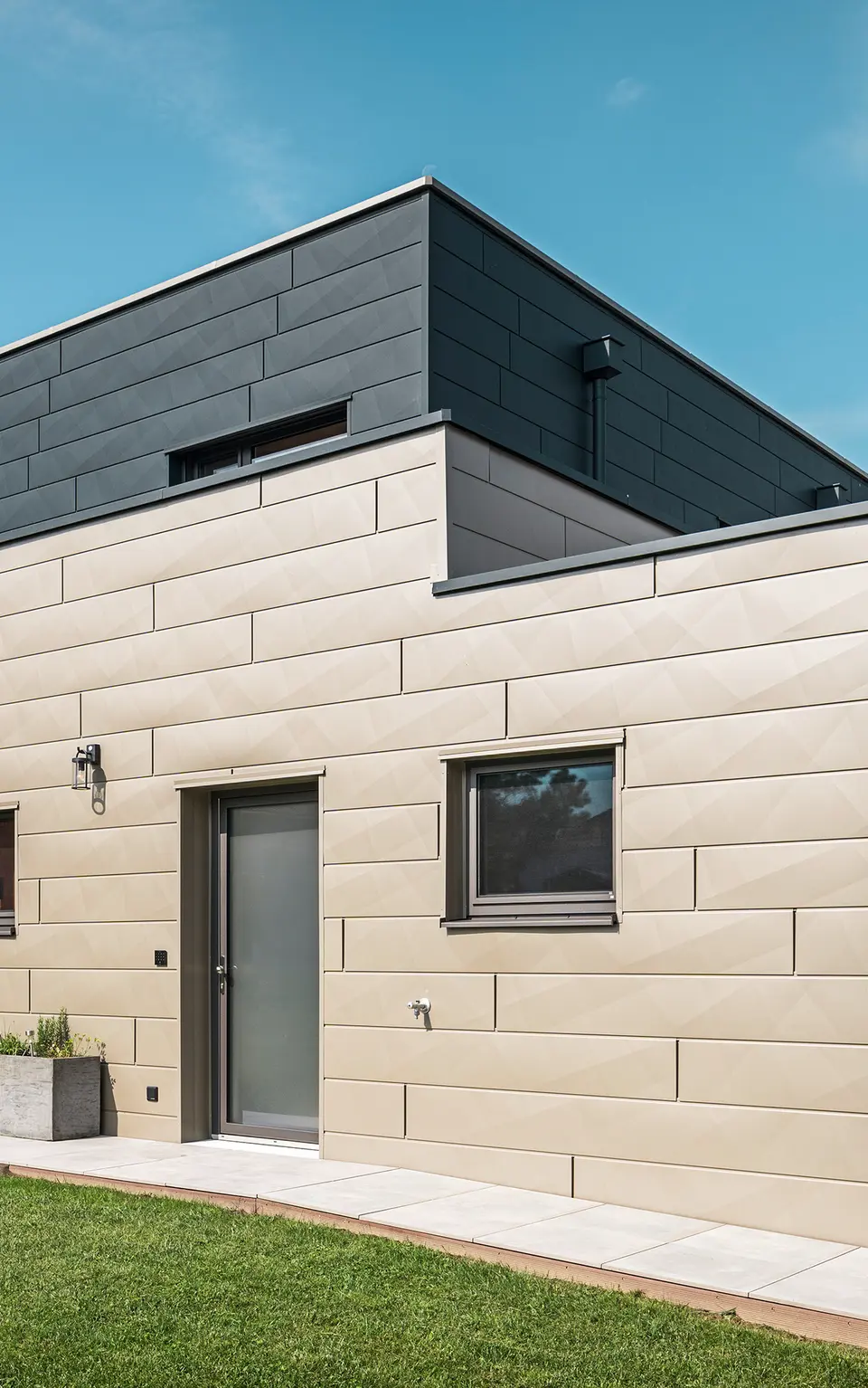Modernes Einfamilienhaus in Wippenham, Österreich, mit zweifarbiger PREFA Aluminiumfassade in Bronze und Anthrazit. Die Fassade ist mit Siding.X Fassadenpaneelen gestaltet, die für eine elegante und zeitgemäße Optik sorgen. Die integrierte PREFA Dachentwässerung mit Wasserfangkasten und Ablaufrohr sowie eine gepflegte Gartenanlage mit Terrasse betonen das moderne Design des Hauses.