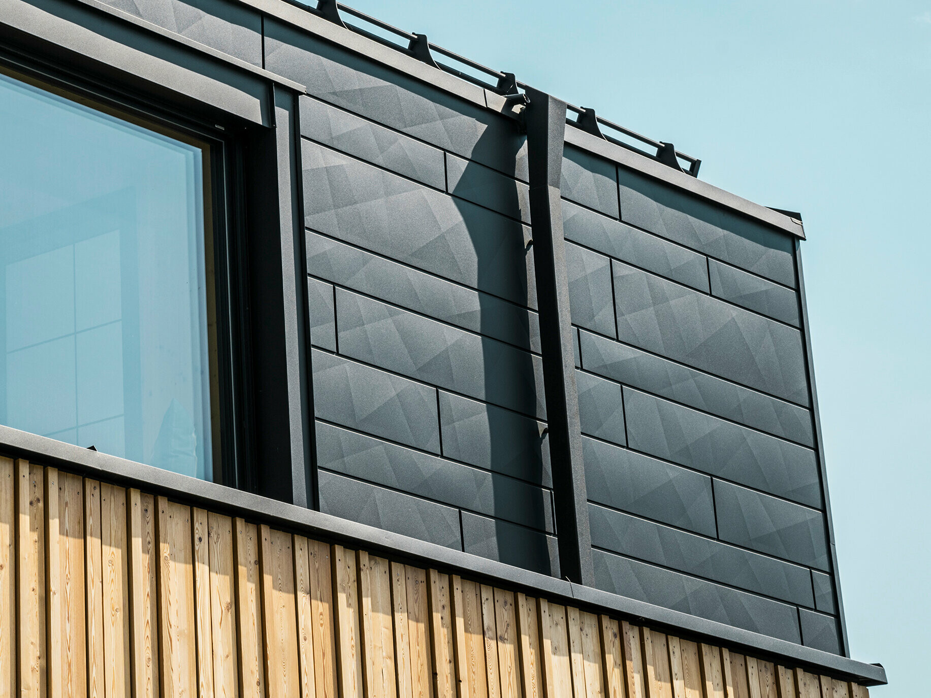 Nahaufnahme des Obergeschosses des Musterhauses Autark im Fertighauszentrum Blaue Lagune, Vösendorf (AT). Die Fassade besteht aus PREFA Siding.X Aluminiumpaneelen in P.10 Schwarz, die durch ihre spezielle Optik mit unterschiedlichen Kantungen auffallen. Darunter befindet sich eine stilvolle Holzverkleidung, die einen harmonischen Kontrast zum dunklen Aluminium bildet. Große Fenster lassen viel Licht in das Gebäudeinnere und betonen das moderne Design.