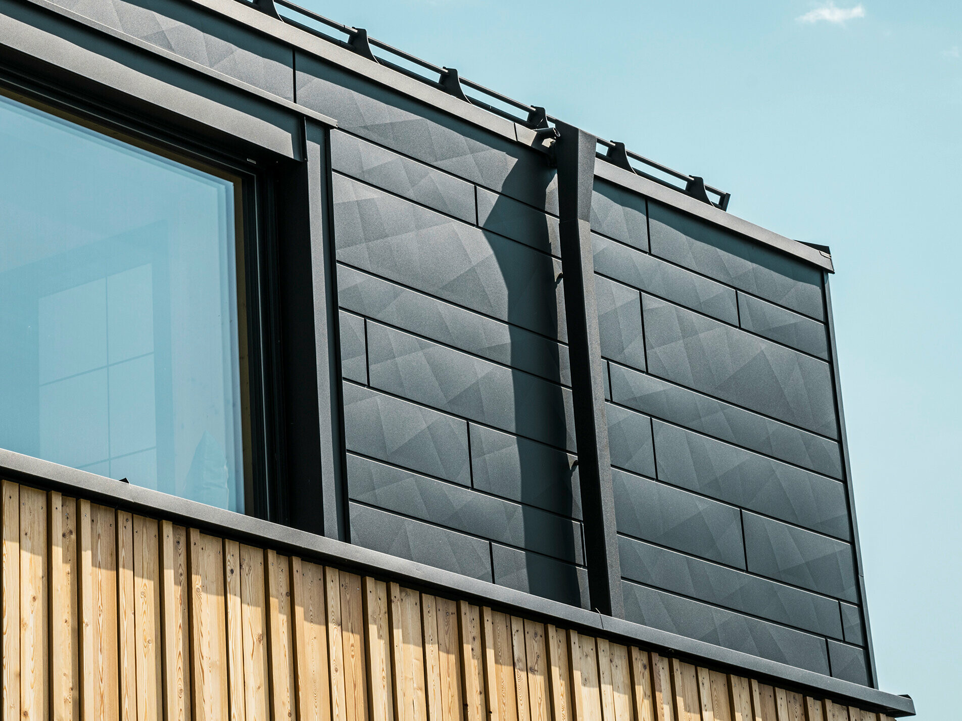 Nahaufnahme des Obergeschosses des Musterhauses Autark im Fertighauszentrum Blaue Lagune, Vösendorf (AT). Die Fassade besteht aus PREFA Siding.X Aluminiumpaneelen in P.10 Schwarz, die durch ihre spezielle Optik mit unterschiedlichen Kantungen auffallen. Darunter befindet sich eine stilvolle Holzverkleidung, die einen harmonischen Kontrast zum dunklen Aluminium bildet. Große Fenster lassen viel Licht in das Gebäudeinnere und betonen das moderne Design.
