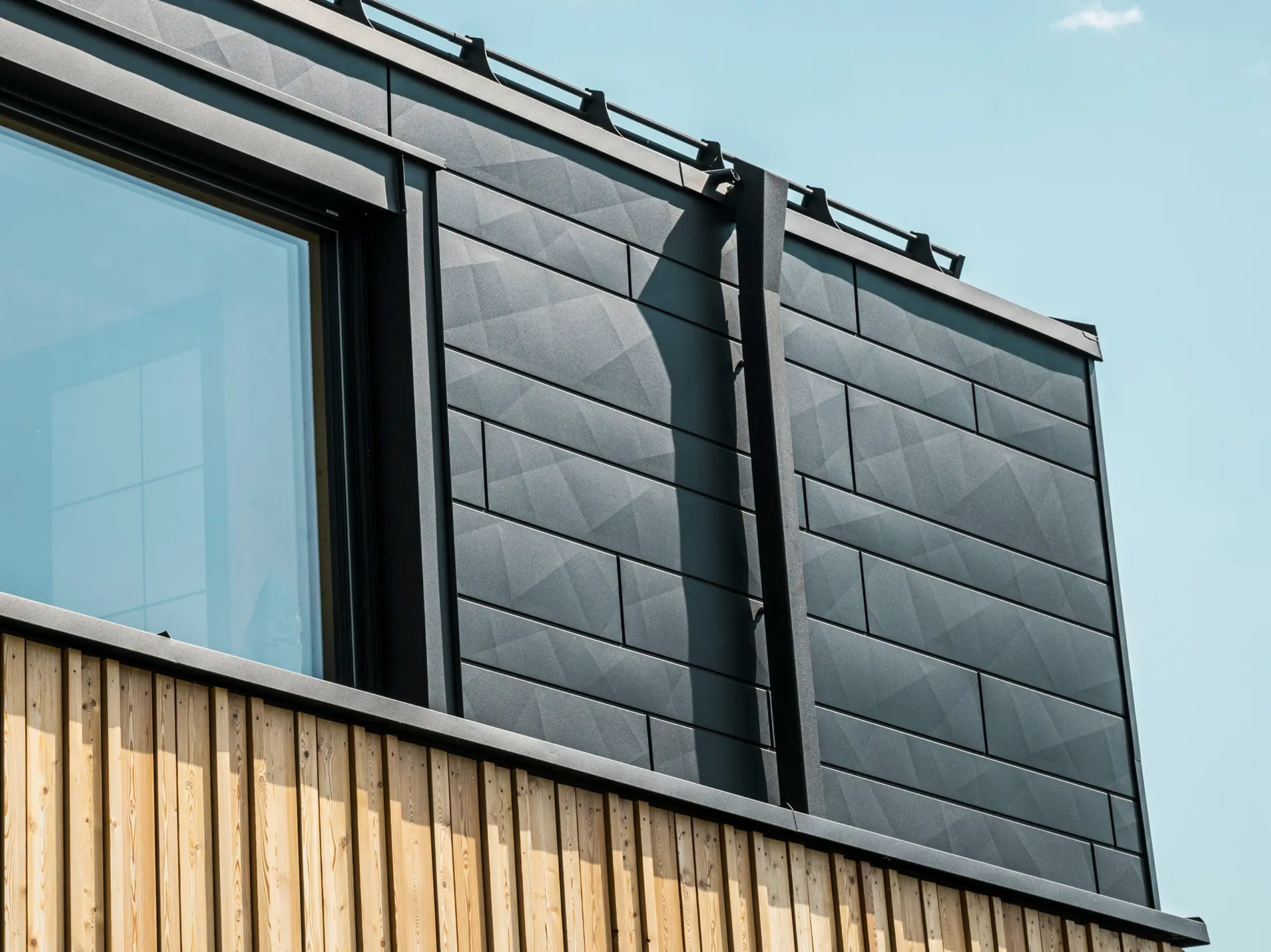 Nahaufnahme des Obergeschosses des Musterhauses Autark im Fertighauszentrum Blaue Lagune, Vösendorf (AT). Die Fassade besteht aus PREFA Siding.X Aluminiumpaneelen in P.10 Schwarz, die durch ihre spezielle Optik mit unterschiedlichen Kantungen auffallen. Darunter befindet sich eine stilvolle Holzverkleidung, die einen harmonischen Kontrast zum dunklen Aluminium bildet. Große Fenster lassen viel Licht in das Gebäudeinnere und betonen das moderne Design.