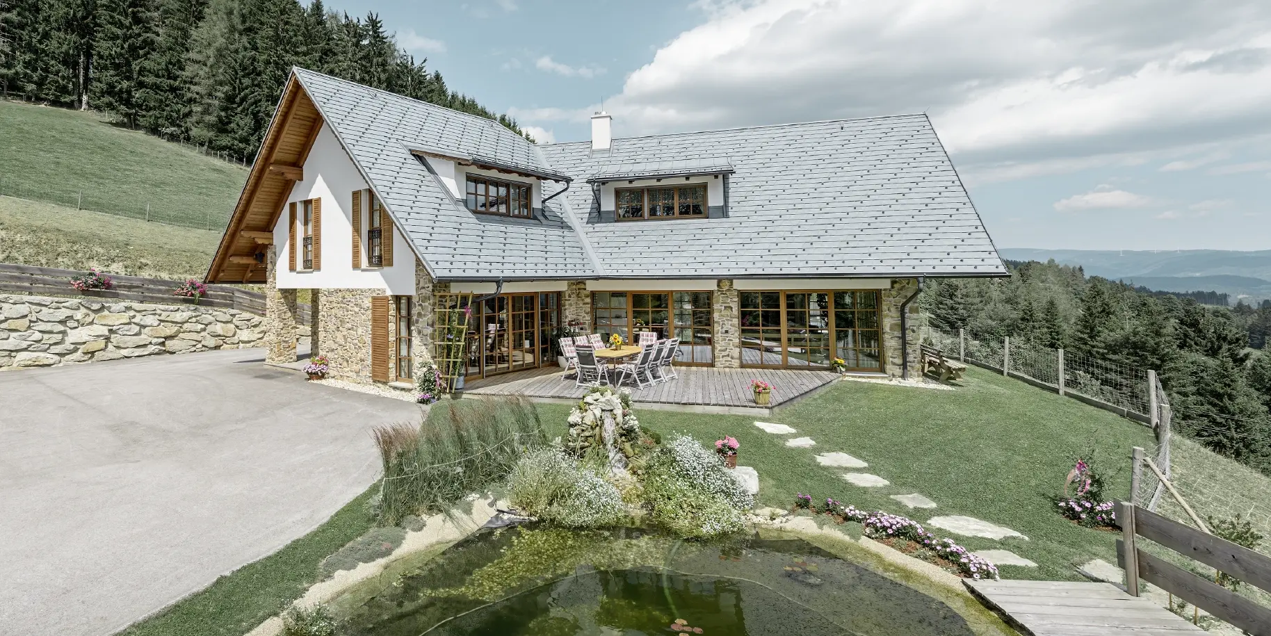 Das Einfamilienhaus in Hanglage, in L-Form gebaut, hat ein Satteldach mit zwei Schleppgauben. Das Dach wurde mit der PREFA Dachschindel in Steingrau eingedeckt. Im Vordergrund ist ein Teich angelegt und im Hintergrund erkennt man einen Wald.