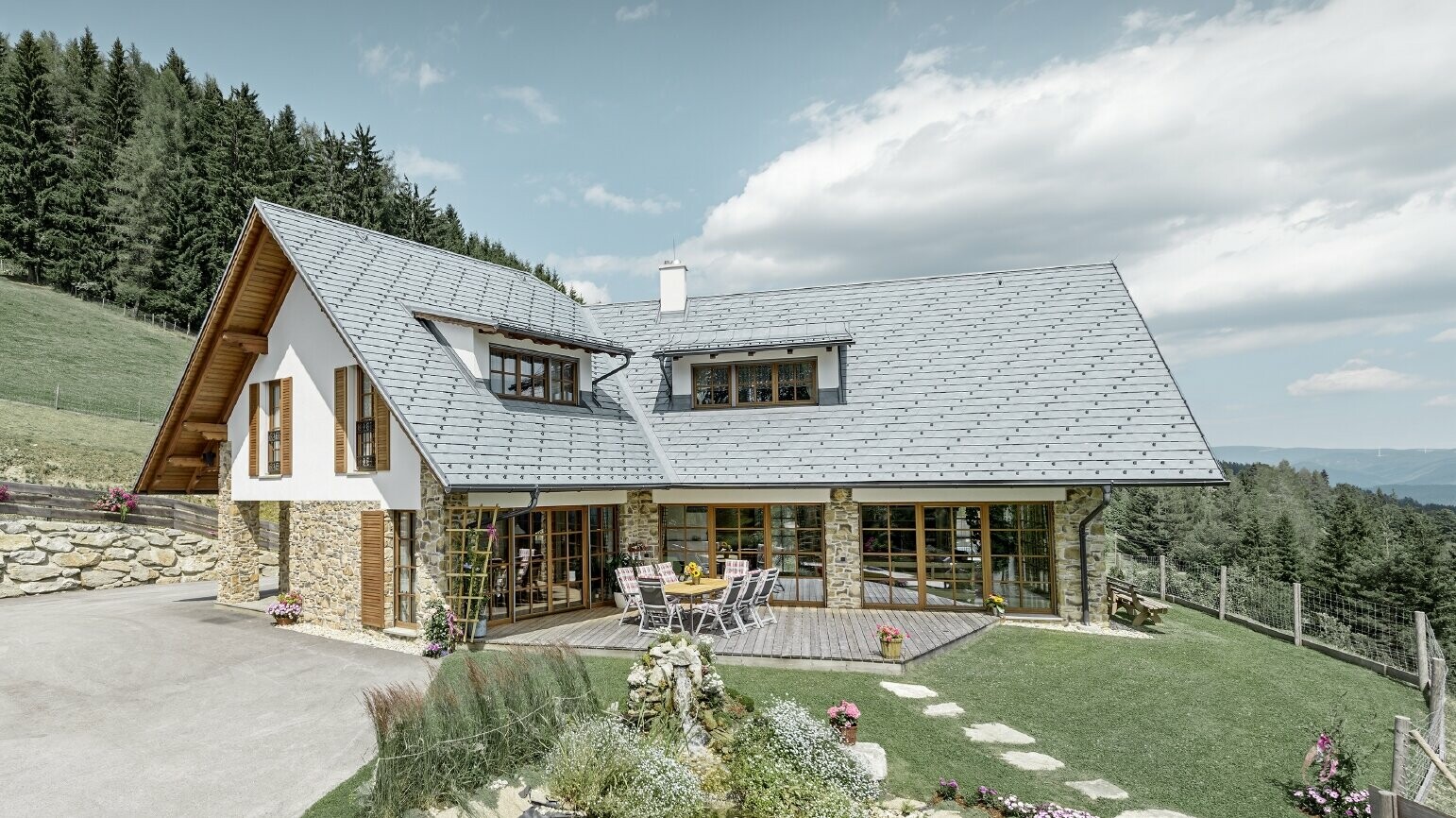 Das Einfamilienhaus in Hanglage, in L-Form gebaut, hat ein Satteldach mit zwei Schleppgauben. Das Dach wurde mit der PREFA Dachschindel in Steingrau eingedeckt. Im Vordergrund ist ein Teich angelegt und im Hintergrund erkennt man einen Wald.