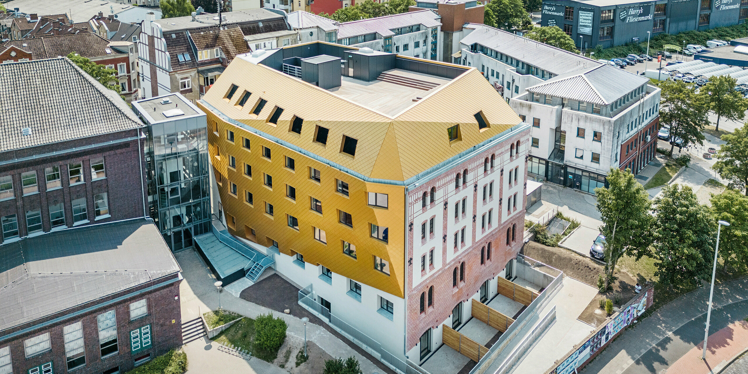 Vogelperspektive auf "The Station" in Kiel. Das Studentenwohnheim ragt im Stadtpanorama hervor durch die markanten Gebäudeteile aus PREFA Dach- und Wandrauten 29 × 29 in edlem Mayagold.