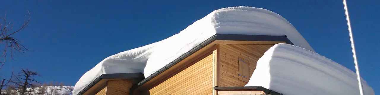 Das Bild zeigt die Hütte Capanna Buffaloraim im Winter, die von dicken Schneewechten am Dach bedeckt ist. Das Bild weist auf die Gefahren durch abrutschende Schneelawinen hin. Das Dach der Berghütte ist mit robusten PREFA Dachpaneelen FX.12-gedeckt, welche besonders für extreme Wetterbedingungen geeignet sind. Trotz der massiven Schneelast bleibt die Dachkonstruktion stabil und sicher. Dies zeigt die hohe Belastbarkeit und Qualität der PREFA Produkte. Die Berghütte selbst besteht aus Holz und fügt sich harmonisch in die winterliche Landschaft ein.