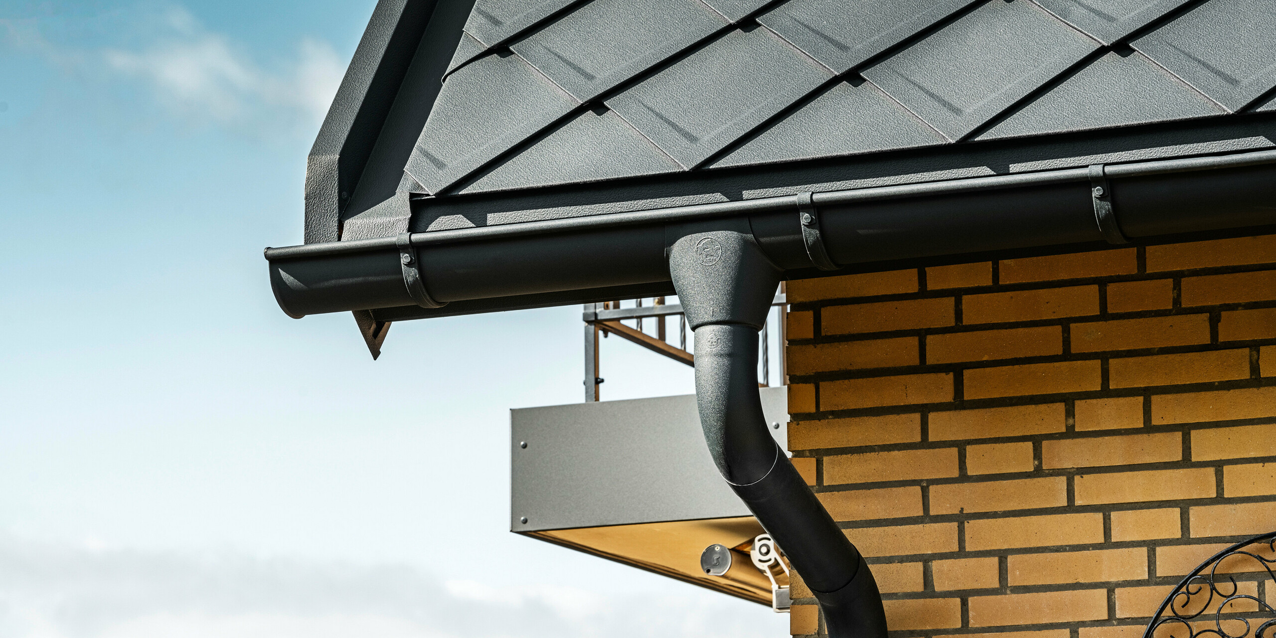 Detailansicht des PREFA Dachentwässerungssystems eines Einfamilienhauses mit anthrazitfarbenen PREFA Dachraute 29×29, welche im Hintergrund am Dach hervorstechen. Zu sehen ist die Hängerinne und das Ablaufrohr in P.10 Anthrazit, die sich nahtlos in das Gesamtdesign einfügen. Die gelben Backsteinwände des Hauses bieten einen natürlichen Farbkontrast zum modernen Dachsystem aus Aluminium.
