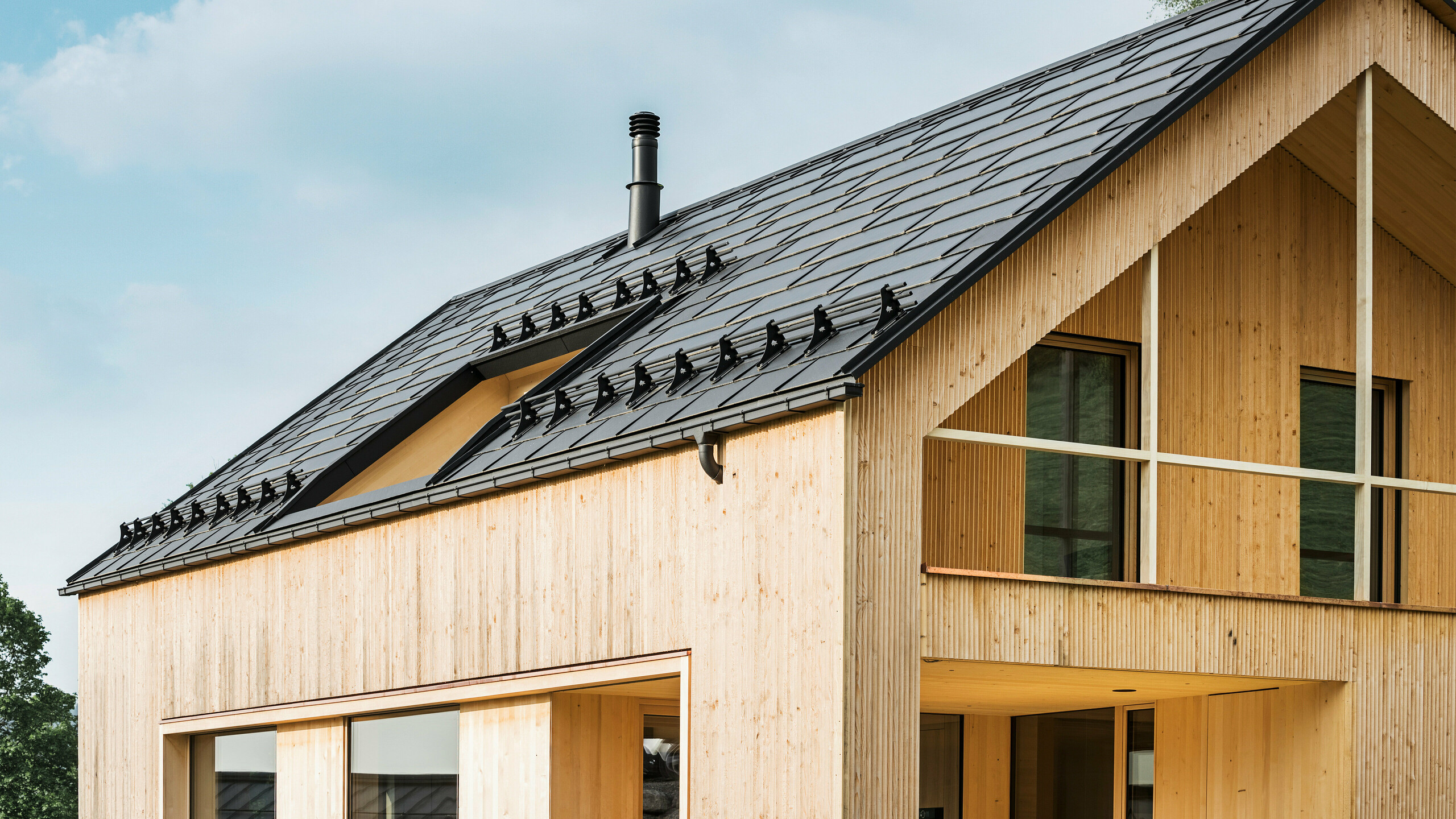 Nahaufnahme des Giebels eines umweltfreundlichen Hauses in Egg, charakterisiert durch PREFA Solardachplatten in elegantem Schwarz, die sich harmonisch in der Mitte der PREFA Dachplatten R.16 einfügen. Die sauberen Linien des Daches kontrastieren mit der natürlichen Textur der Holzfassade, während die Solarmodule ein Statement für nachhaltiges Wohnen setzen. Der subtile Schornstein fügt sich in das Gesamtbild ein, das umweltbewusstes Design mit ländlicher Ästhetik vereint. Zudem verfügt das innovative Ökohaus über zwei Balkone, die einen Blick in die traumhafte Bergkulisse von Egg in Vorarlberg ermöglichen.
