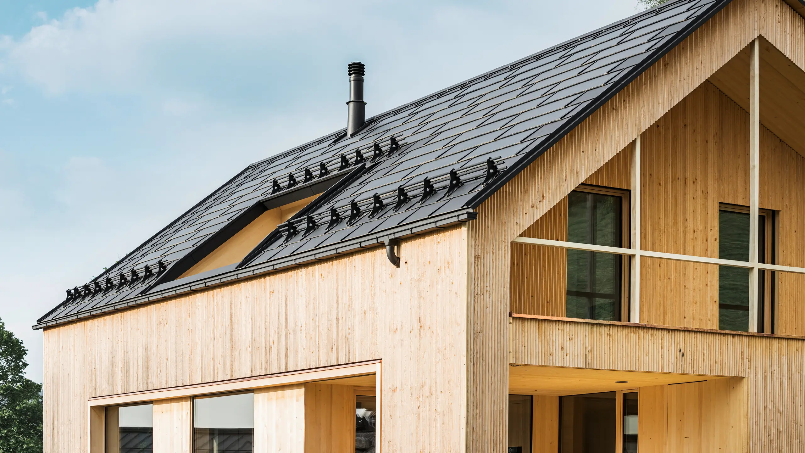 Nahaufnahme des Giebels eines umweltfreundlichen Hauses in Egg, charakterisiert durch PREFA Solardachplatten in elegantem Schwarz, die sich harmonisch in der Mitte der PREFA Dachplatten R.16 einfügen. Die sauberen Linien des Daches kontrastieren mit der natürlichen Textur der Holzfassade, während die Solarmodule ein Statement für nachhaltiges Wohnen setzen. Der subtile Schornstein fügt sich in das Gesamtbild ein, das umweltbewusstes Design mit ländlicher Ästhetik vereint. Zudem verfügt das innovative Ökohaus über zwei Balkone, die einen Blick in die traumhafte Bergkulisse von Egg in Vorarlberg ermöglichen.