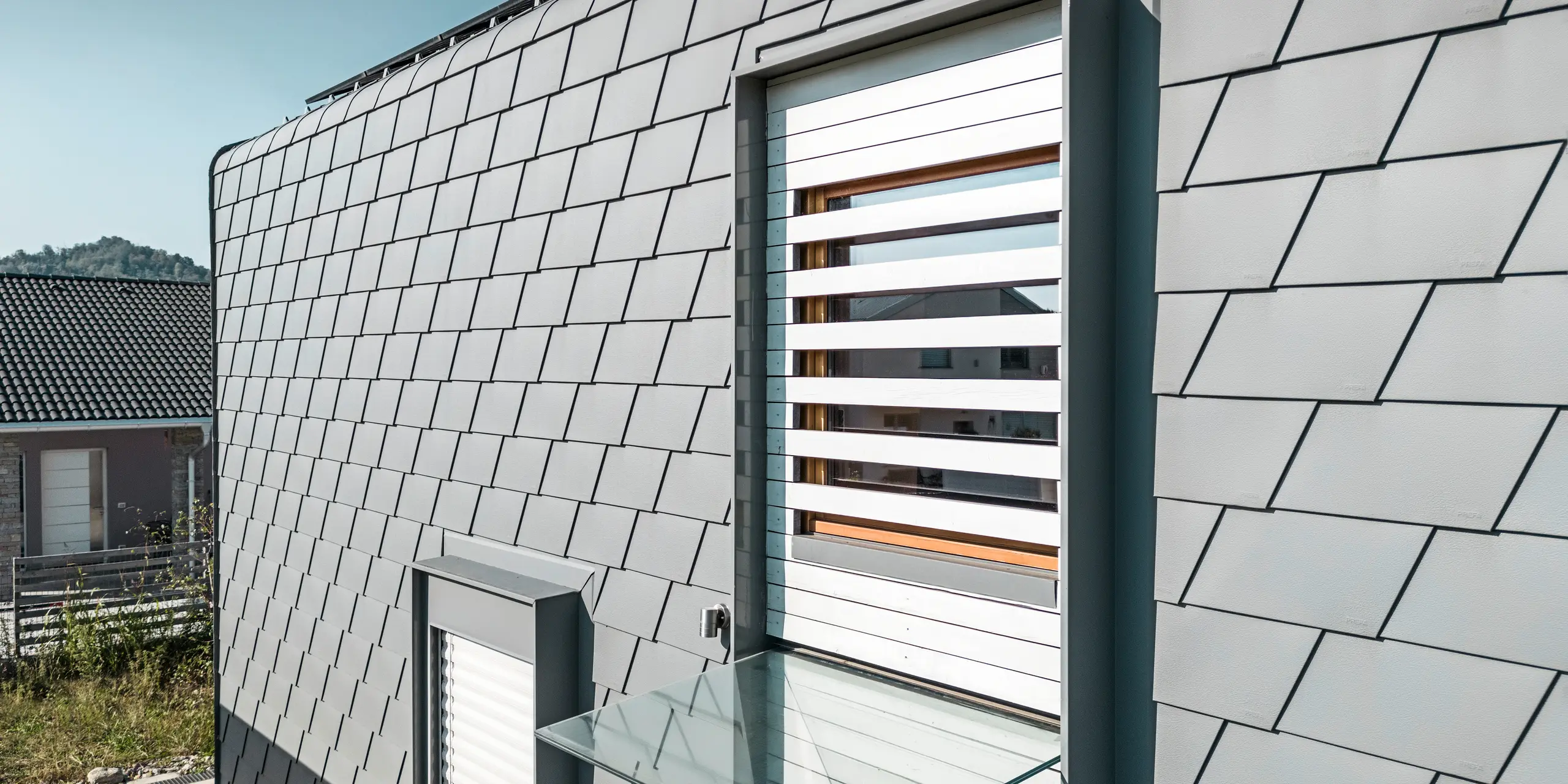 Detailaufnahme eines Fensters mit weißen Außenjalousien. Die Fassade des modernen Hauses ist mit PREFA Wandschindeln in der Farbe P.10 Hellgrau verkleidet. Das Dach ist mit PREFA Dachschindeln in P.10 Hellgrau eingedeckt. Die klare Geometrie der Schindeln ergibt eine strukturierte Oberfläche, die durch die natürlichen Schattenwürfe zusätzlich betont wird. Das Fenster mit den Außenjalousien durchbricht das Muster und fügt ein warmes, natürliches Element in das kühle Grau der Schindeln ein.
