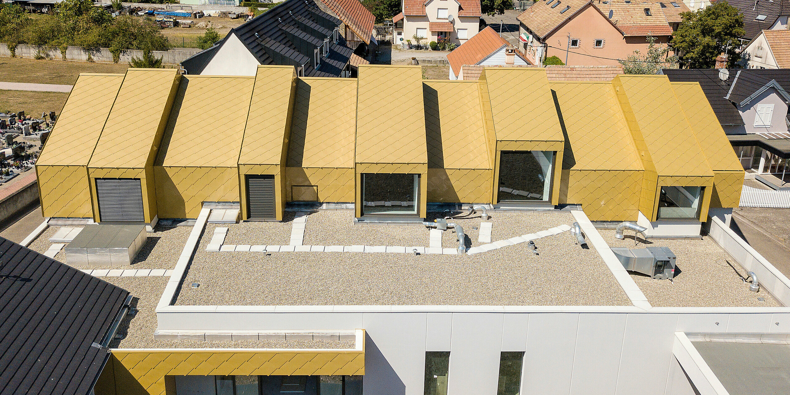 Vogelperspektive auf das Dach des Gemeinde- und Kulturzentrums Fessenheim, Frankreich. Das Dach sowie Teile der Fassade wurden mit PREFA Dach- und Wandraute 44 × 44 in Sparkling Gold verkleidet.