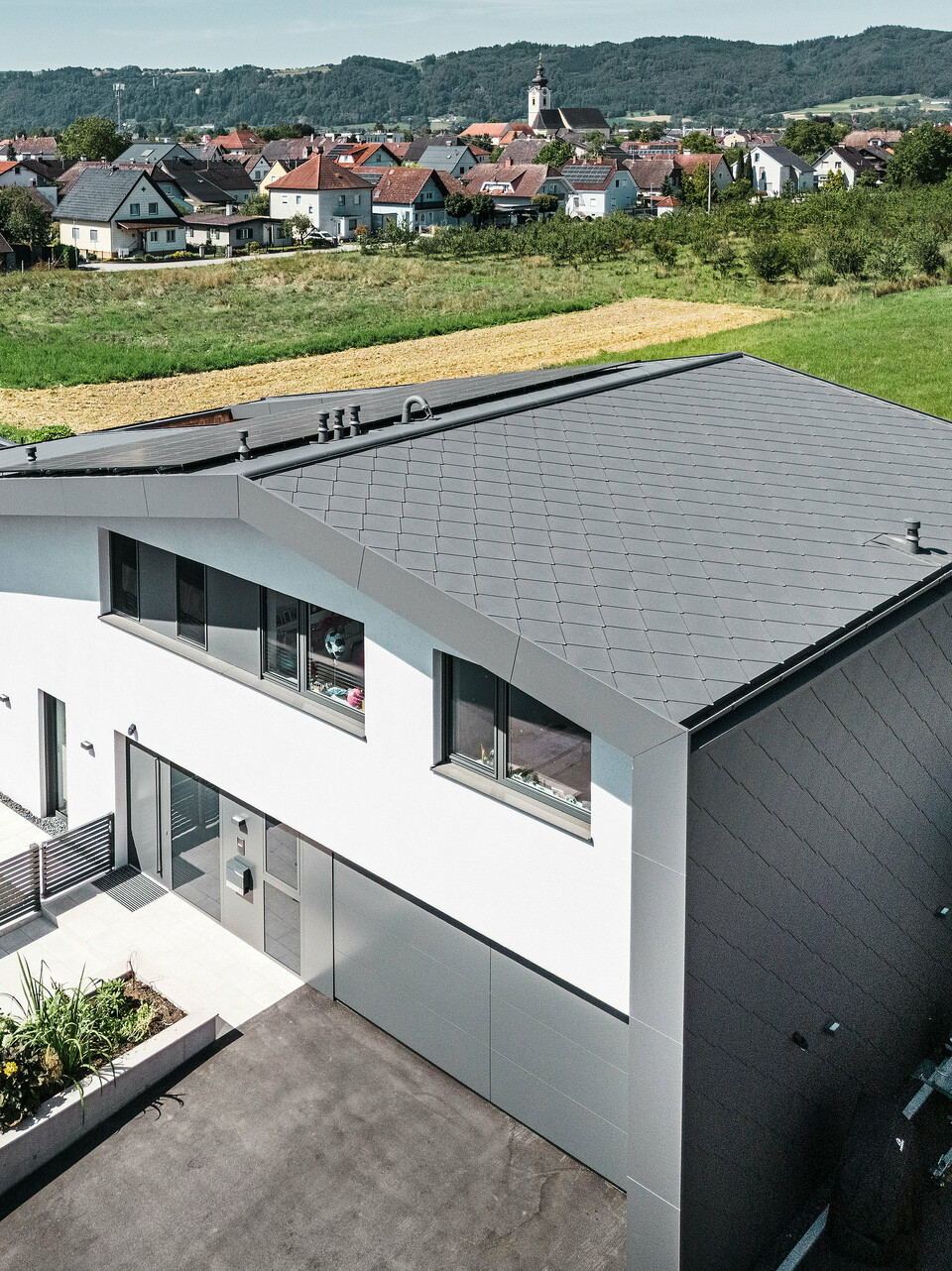 Luftaufnahme eines modernen Einfamilienhauses in Aschach mit zeitloser Architektur. Fassade und Dach sind mit dunkelgrauen PREFA Aluminium-Rauten 44 × 44 eingedeckt. Die langlebige und witterungsbeständige Hülle verleiht dem Gebäude ein elegantes Aussehen und bietet optimalen Schutz vor Umwelteinflüssen. Vor dem Haus befindet sich ein gepflegter Vorgarten mit einer saftig grünen Wiese. Rechts vom Haus liegt ein kleines Nebengebäude und neben der Eingangstür befindet sich eine großzügige Garage mit asphaltierter Zufahrt.