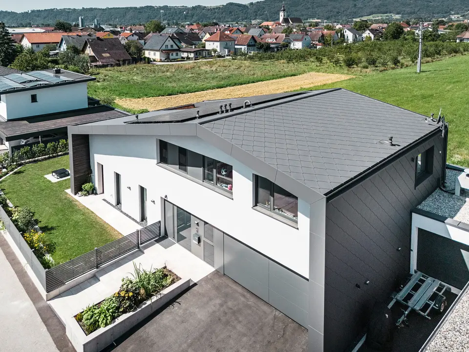 Luftaufnahme eines modernen Einfamilienhauses in Aschach mit zeitloser Architektur. Fassade und Dach sind mit dunkelgrauen PREFA Aluminium-Rauten 44 × 44 eingedeckt. Die langlebige und witterungsbeständige Hülle verleiht dem Gebäude ein elegantes Aussehen und bietet optimalen Schutz vor Umwelteinflüssen. Vor dem Haus befindet sich ein gepflegter Vorgarten mit einer saftig grünen Wiese. Rechts vom Haus liegt ein kleines Nebengebäude und neben der Eingangstür befindet sich eine großzügige Garage mit asphaltierter Zufahrt.