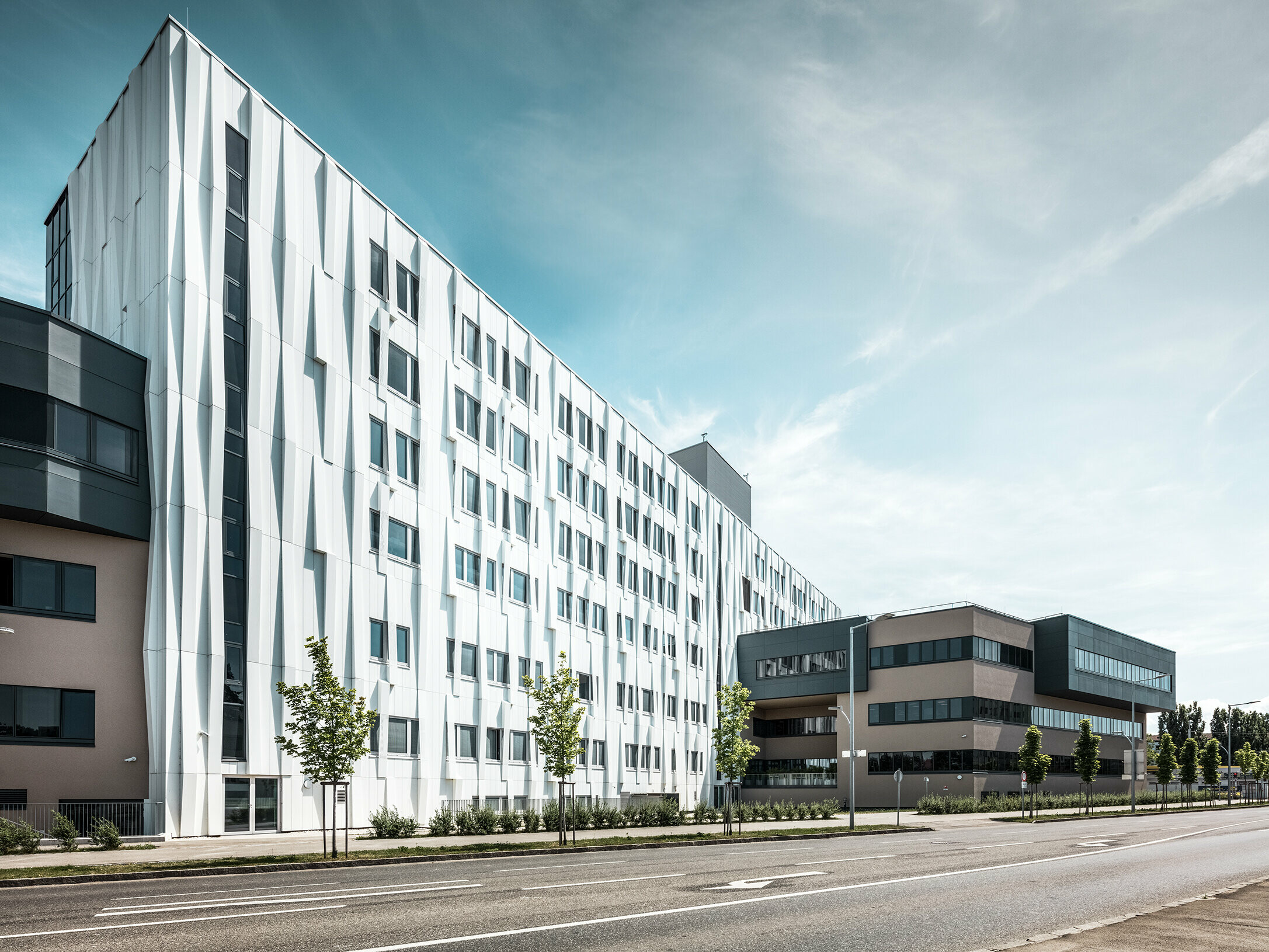 Seitliche Ansicht des Universitätsklinikums in St. Pölten, Niederösterreich. Die robuste Aluminiumfassade macht das Gebäude zu einem Blickfang in der Stadt. Im Vordergrund ist eine Straße mit einigen Bäumen zu sehen.