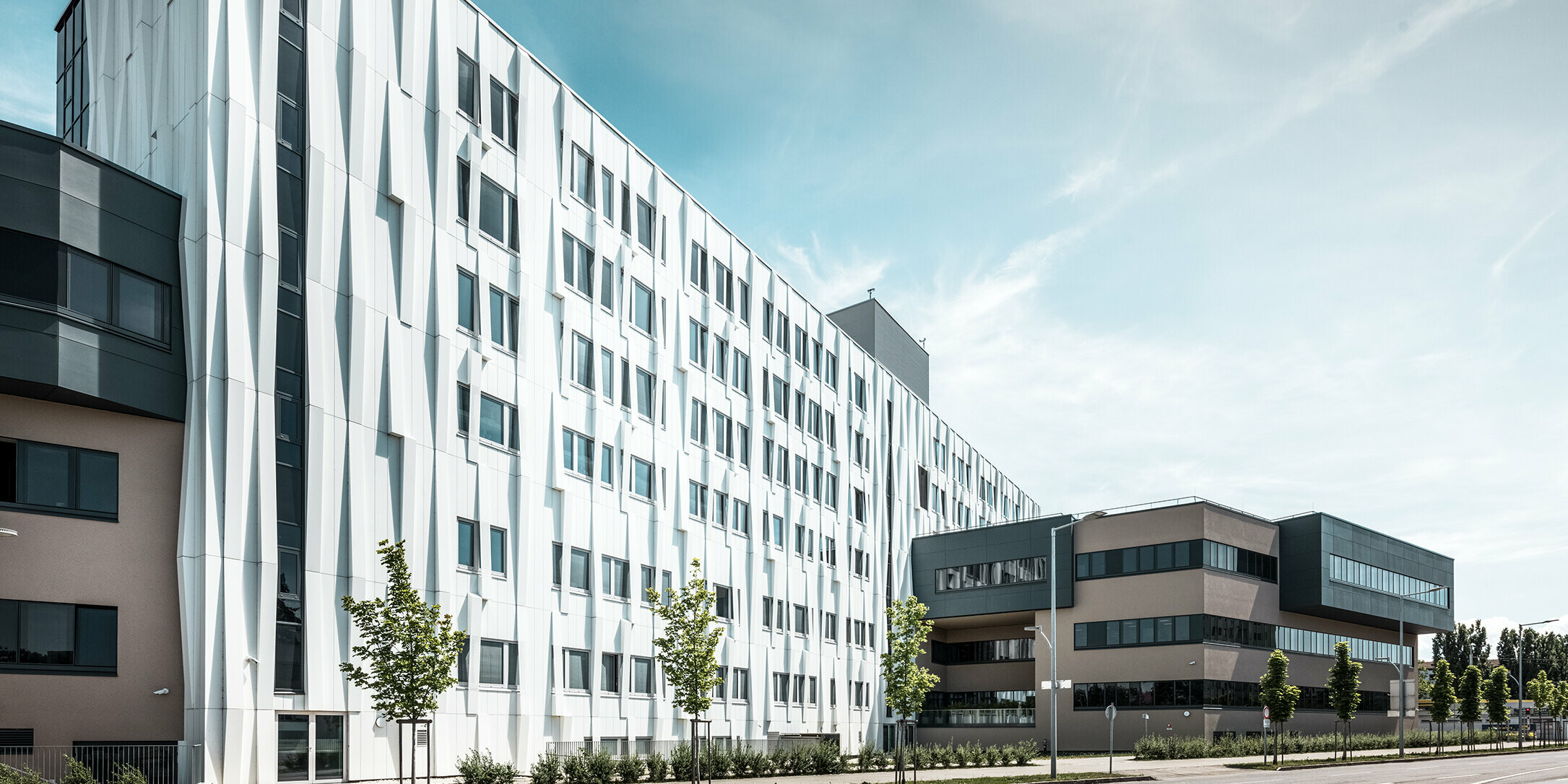 Seitliche Ansicht des Universitätsklinikums in St. Pölten, Niederösterreich. Die robuste Aluminiumfassade macht das Gebäude zu einem Blickfang in der Stadt. Im Vordergrund ist eine Straße mit einigen Bäumen zu sehen.