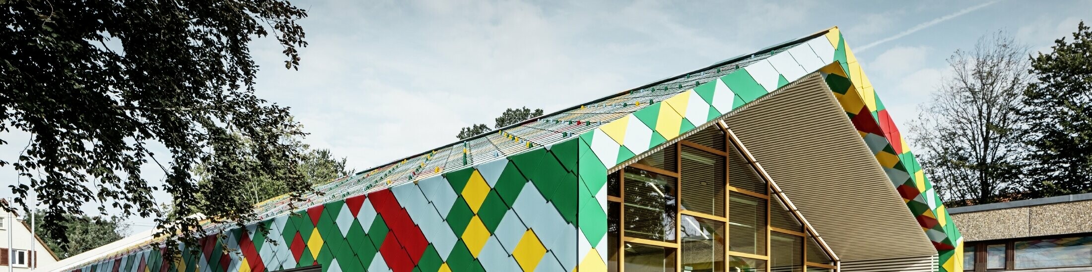 Modernes Gebäude mit auffälliger Fassade aus bunten Aluminiumrauten von PREFA in Grün, Rot, Gelb und Hellblau. Die Dach- und Wandflächen gehen fließend ineinander über und bilden ein geometrisches Muster. Der Eingangsbereich besteht aus großen Glasfronten mit Holzrahmen. Das Gebäude steht in einer grünen Umgebung mit Bäumen und weiteren Schul- oder Verwaltungsgebäuden im Hintergrund.