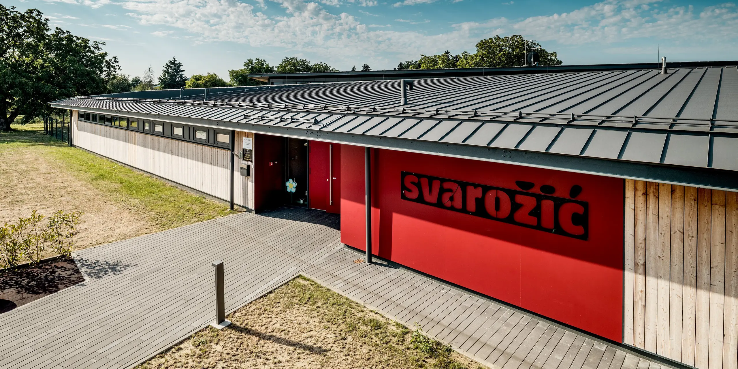 Außenansicht des Kindergartens Svarozic mit moderner Holzfassade, markantem roten Eingangsbereich und großflächigem PREFALZ Aluminiumdach in P.10 Anthrazit. Die klare Stehfalzstruktur unterstreicht die reduzierte Architektur und sorgt für einen langlebigen, widerstandsfähigen Witterungsschutz. PREFA Aluminium verbindet Funktionalität und zeitlose Ästhetik in einer harmonischen Gebäudegestaltung.