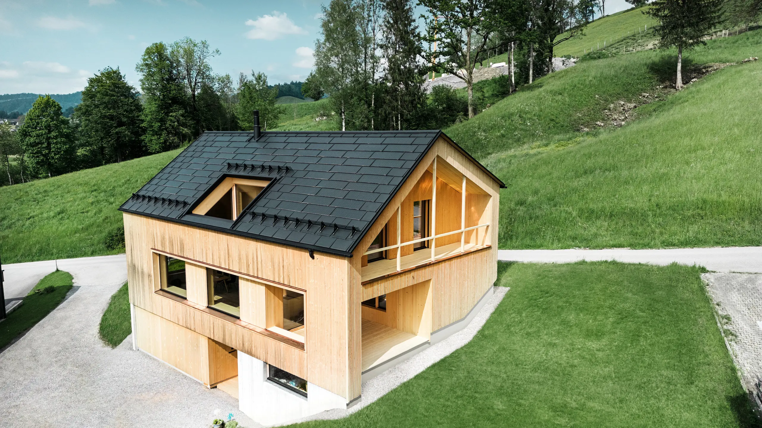 Das Bild präsentiert ein malerisches Ökohaus in Egg, eingebettet in die sanften grünen Hügel, mit einem nachhaltigen PREFA Solardach. Die schwarzen, hochqualitativen PREFA Dachplatten R.16 sowie die PREFA Solardachplatten fügen sich harmonisch in die ländliche Landschaft und betonen die natürliche Schönheit der Umgebung. Die Solardachplatten sind nicht nur ein Bekenntnis zu erneuerbarer Energie, sondern auch ein Designelement, das die moderne Holzfassade und die klaren Linien des Gebäudes stilvoll ergänzt. Dieses Haus ist ein Vorbild für umweltbewusstes Bauen und eine Inspiration für zukunftsorientierte Architektur.