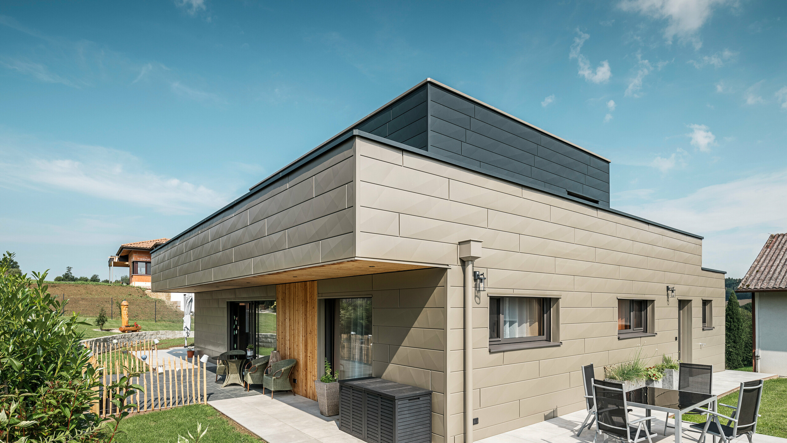 Ein modernes Einfamilienhaus in Wippenham, Österreich, mit einer zweifarbigen PREFA Aluminiumfassade, die aus den extravaganten Siding.X Fassadenpaneelen in Bronze und Anthrazit besteht. Die Fassade zeigt klar erkennbare, elegante Linien und Kantungen. Deutlich sichtbar ist die harmonisch integrierte PREFA Dachentwässerung mit Wasserfangkasten und Ablaufrohr, die sich nahtlos in das architektonische Gesamtkonzept einfügt. Im Vordergrund ist eine gemütliche Terrasse mit Gartenmöbeln zu sehen, die das moderne Design des neugebauten Hauses unterstreicht.